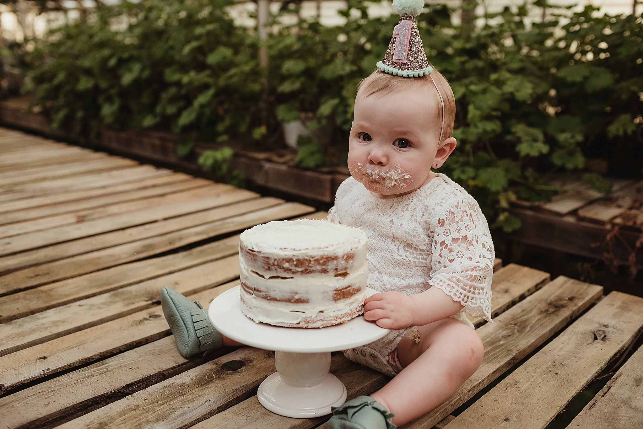 Baby in lace outfit sits with naked birthday cake on wooden deck.