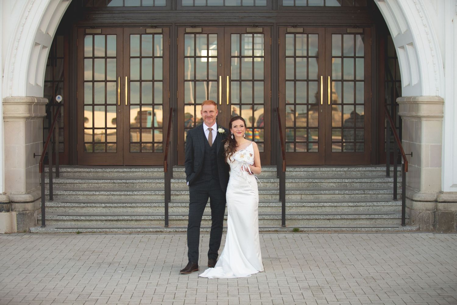 Couple in formal wedding attire poses in front of grand arched entrance.