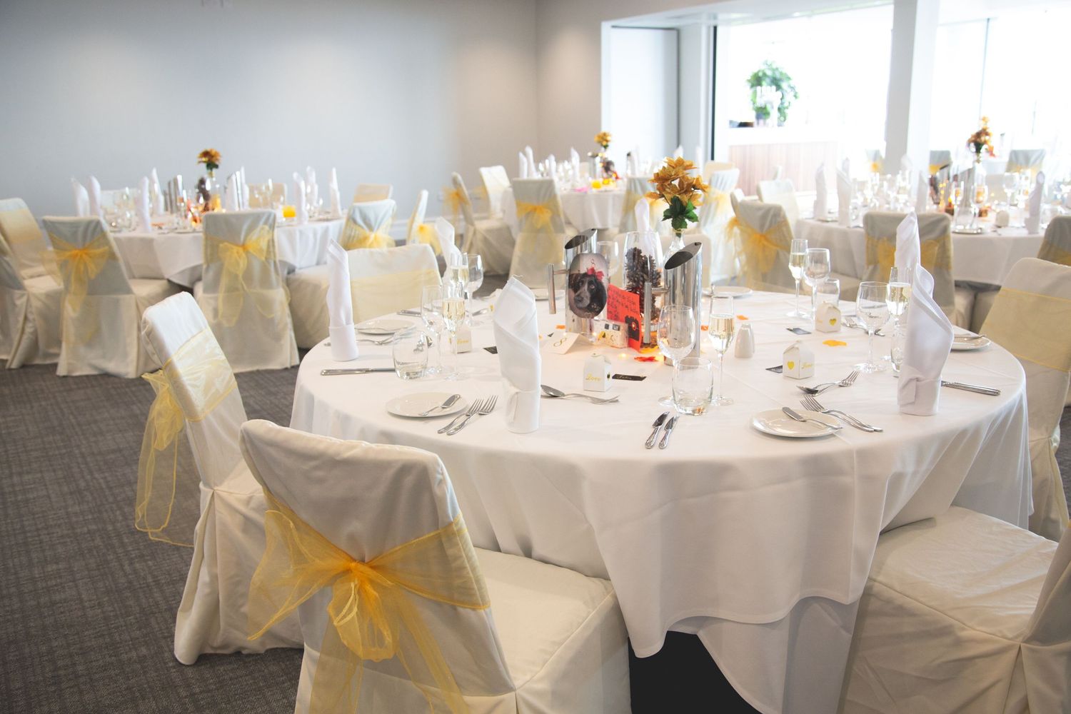 Elegant wedding reception tables with white linens and yellow chair sashes in bright room.
