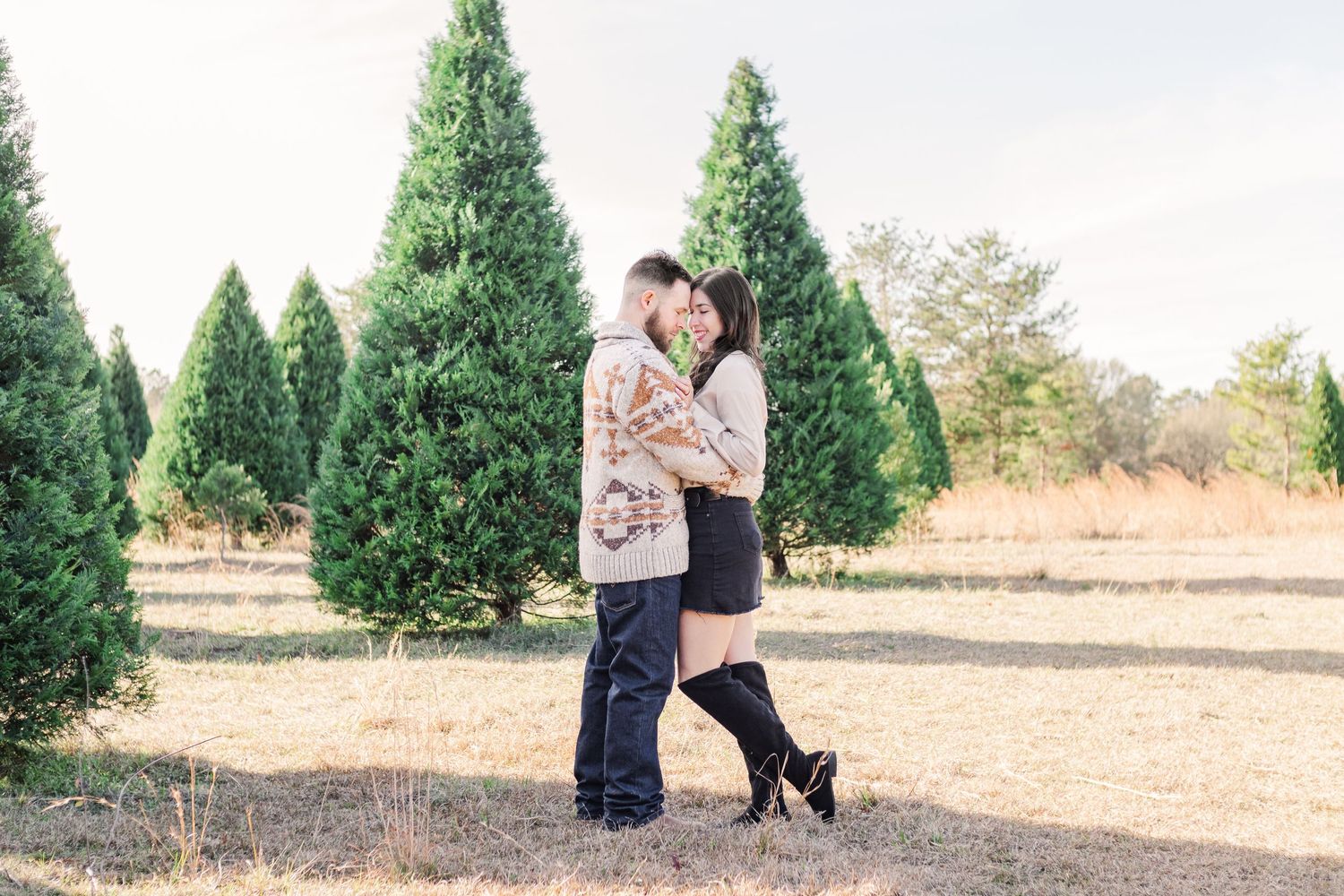 Couple embracing in romantic moment among evergreen trees at outdoor photo session.