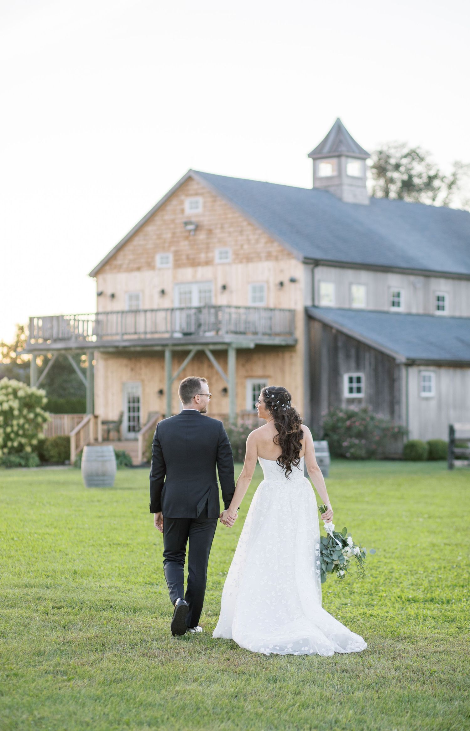 Wedding couple walks toward rustic barn venue with classic architecture and cupola.