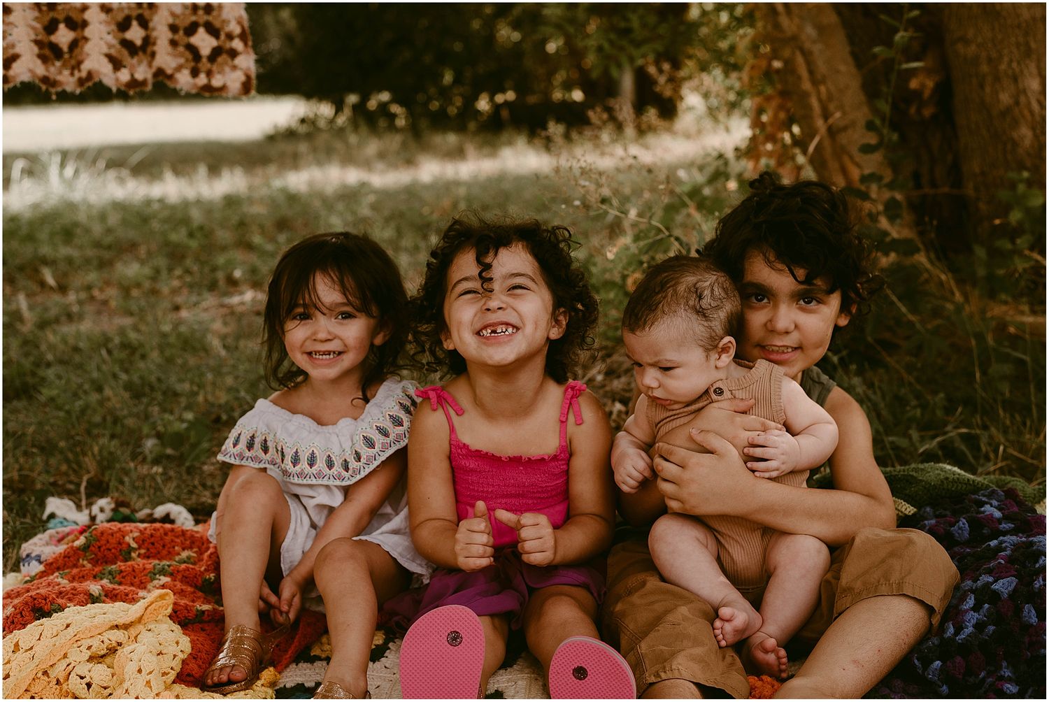 Children sitting together outdoors on a blanket with retro-patterned backdrop on a sunny day.