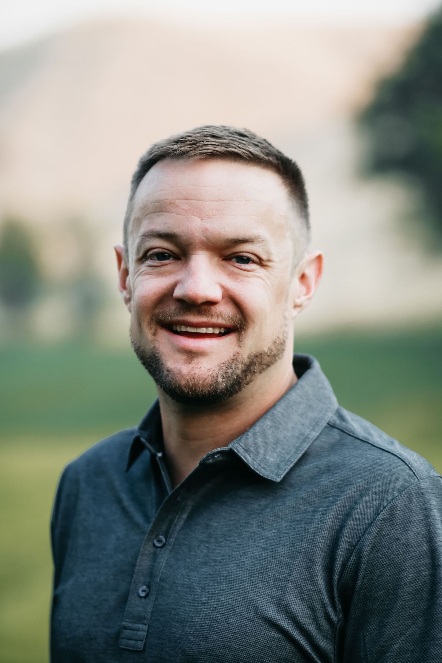 A man in a grey collared shirt smiles, professional headshot outdoors