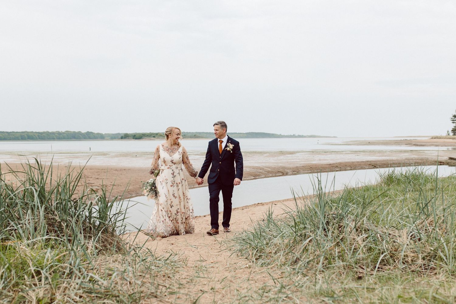 Couple taking romantic beach photos with dune grass and shoreline in the background.