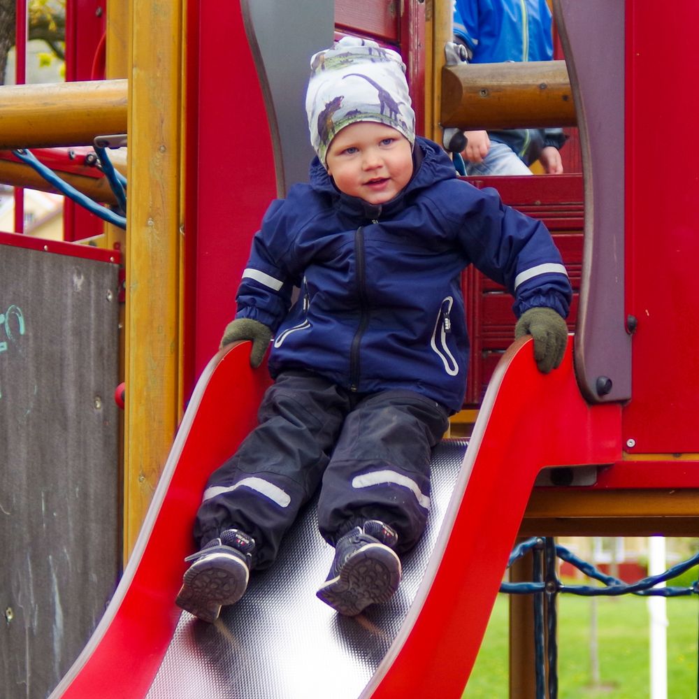 A youngster in a navy snowsuit slides down a red playground slide.
