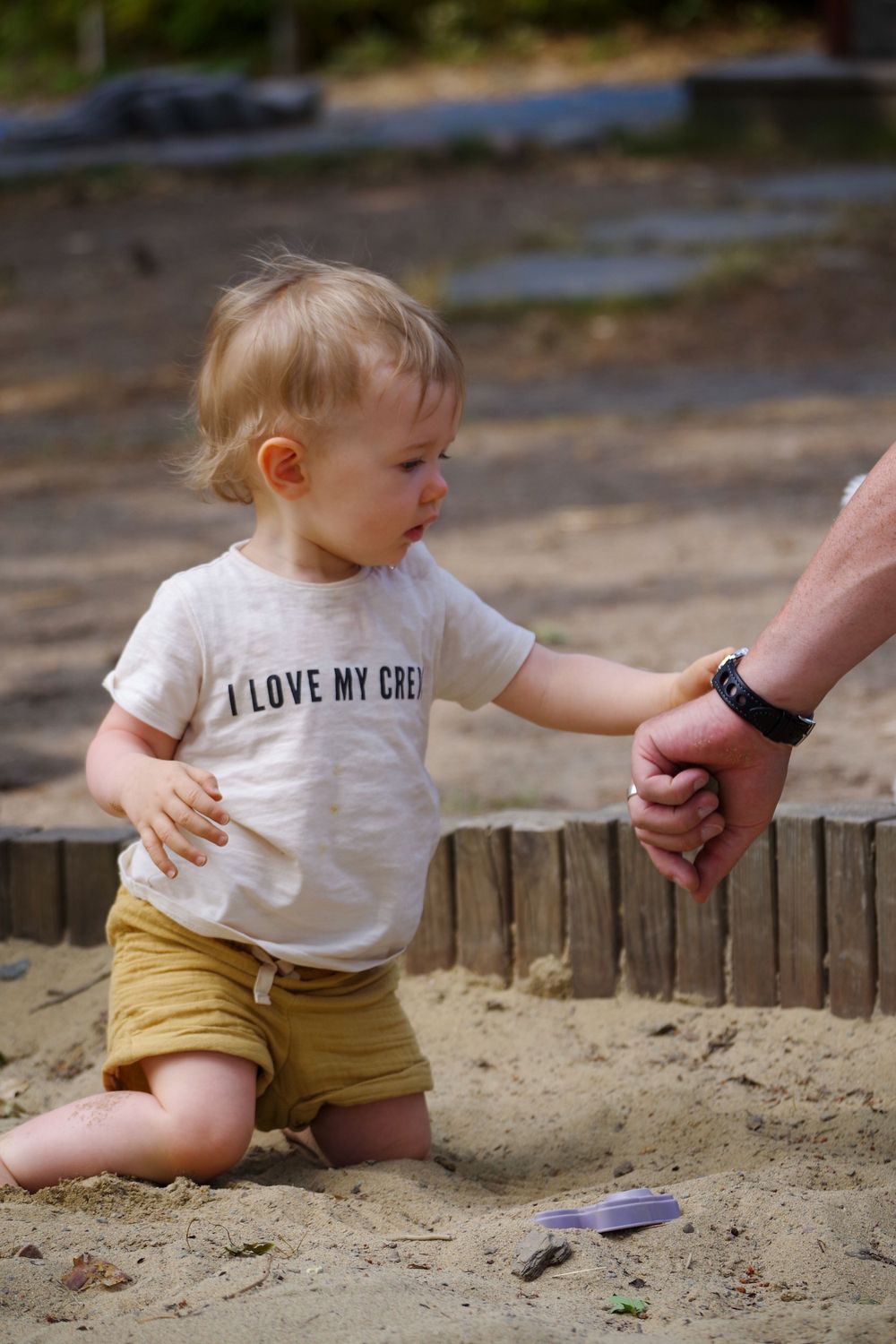 A small child wearing a white t-shirt and yellow shorts plays in a sandbox while holding an adult's hand.