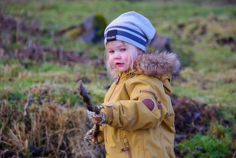 A child in a mustard-colored winter coat and blue striped hat explores outdoors with a stick.