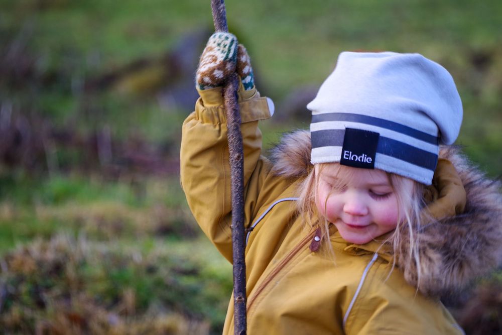 Someone in a yellow coat and striped beanie plays outdoors while holding a stick.