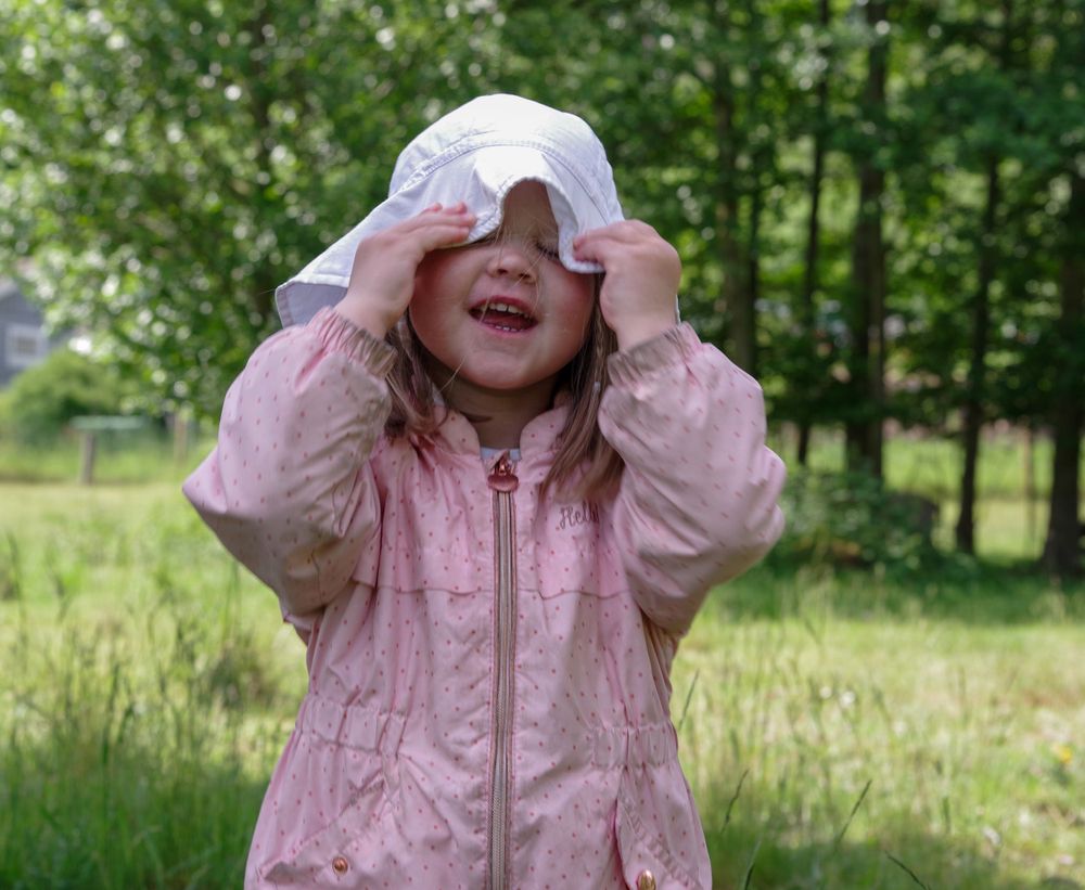 Small child wearing pink jacket and white hat plays playfully in a grassy outdoor setting.