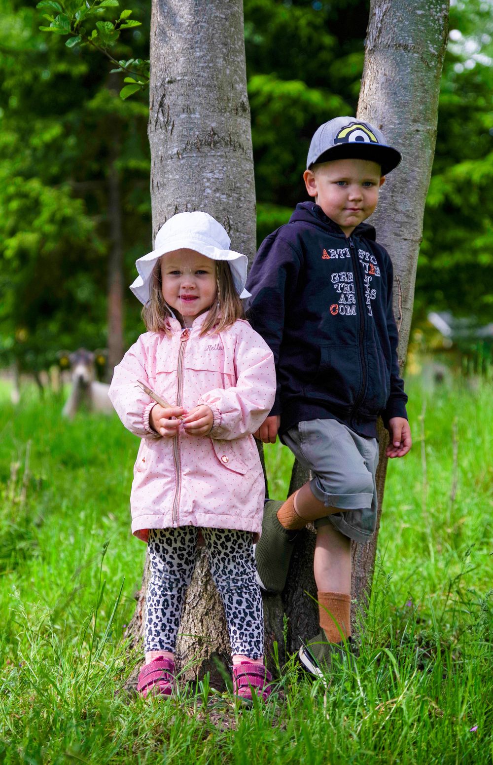 Two children stand by a tree trunk in a grassy field wearing casual outdoor clothing.
