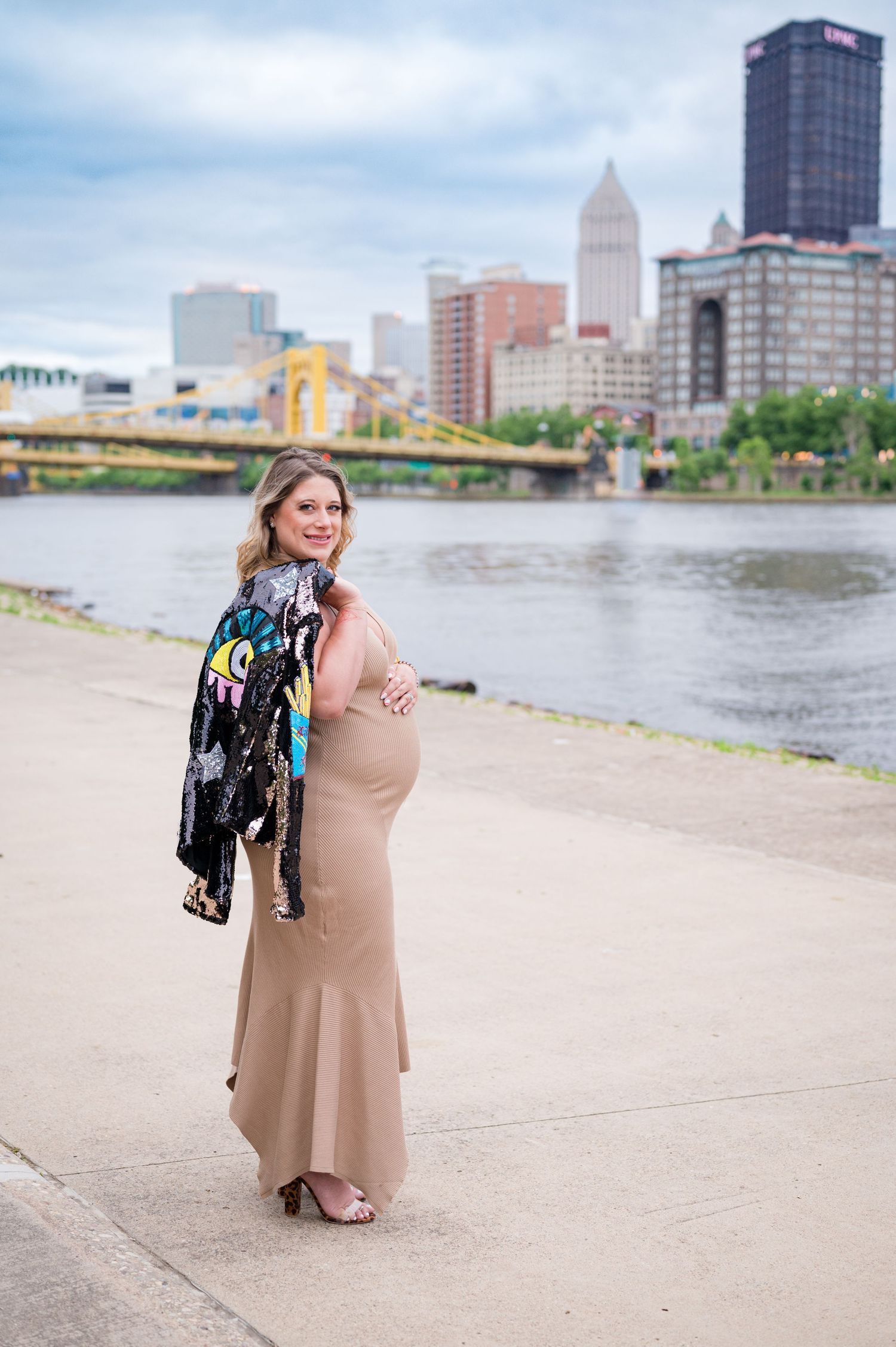 A pregnant mother in a beige dress poses by a river in Pittsburgh with the city skyline in the background.
