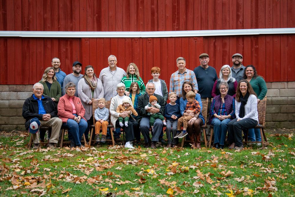 Large group of people sitting together in front of a red barn during autumn.