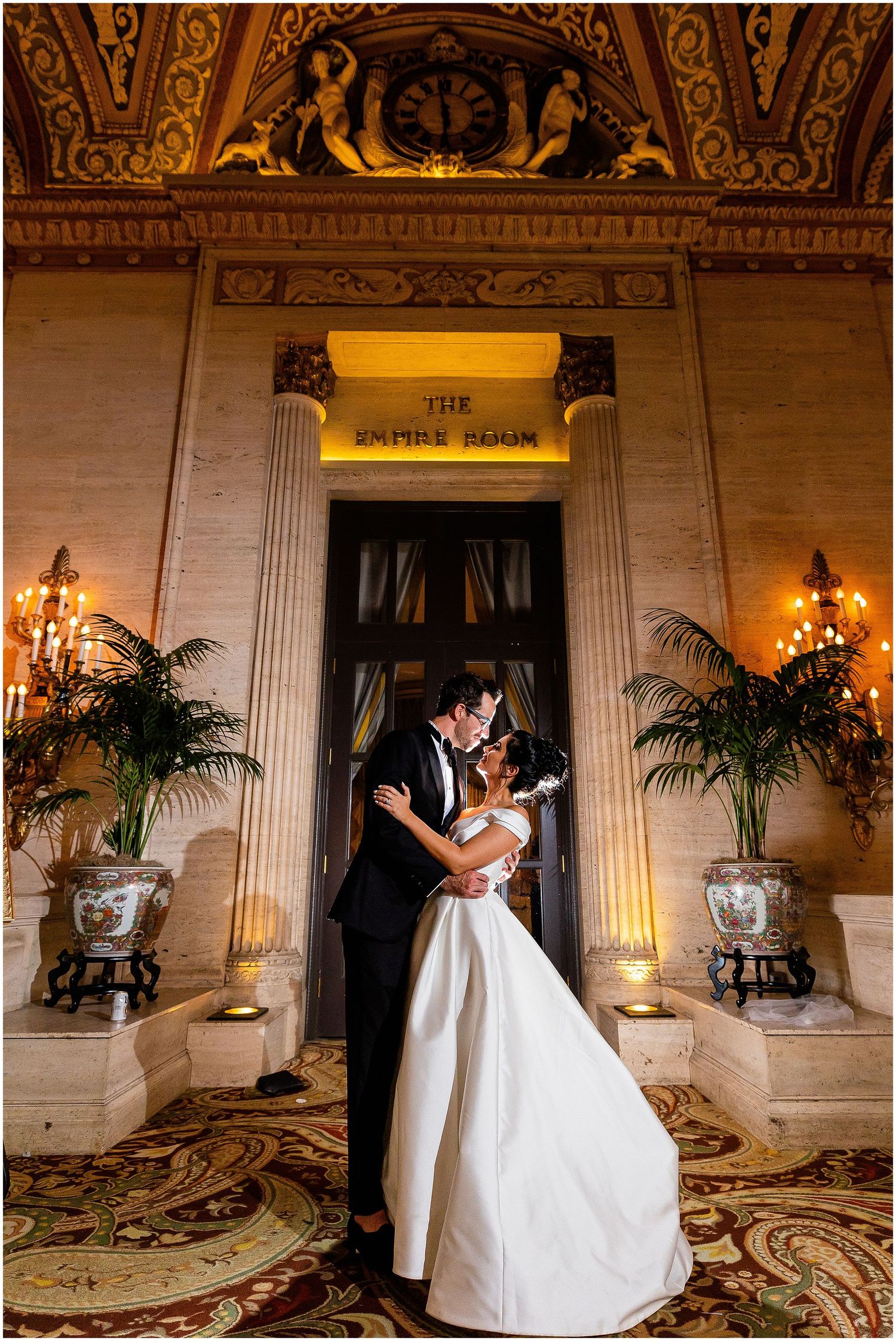 An elegant wedding photo captured in a grand marble entrance hall with ornate decor and warm lighting.