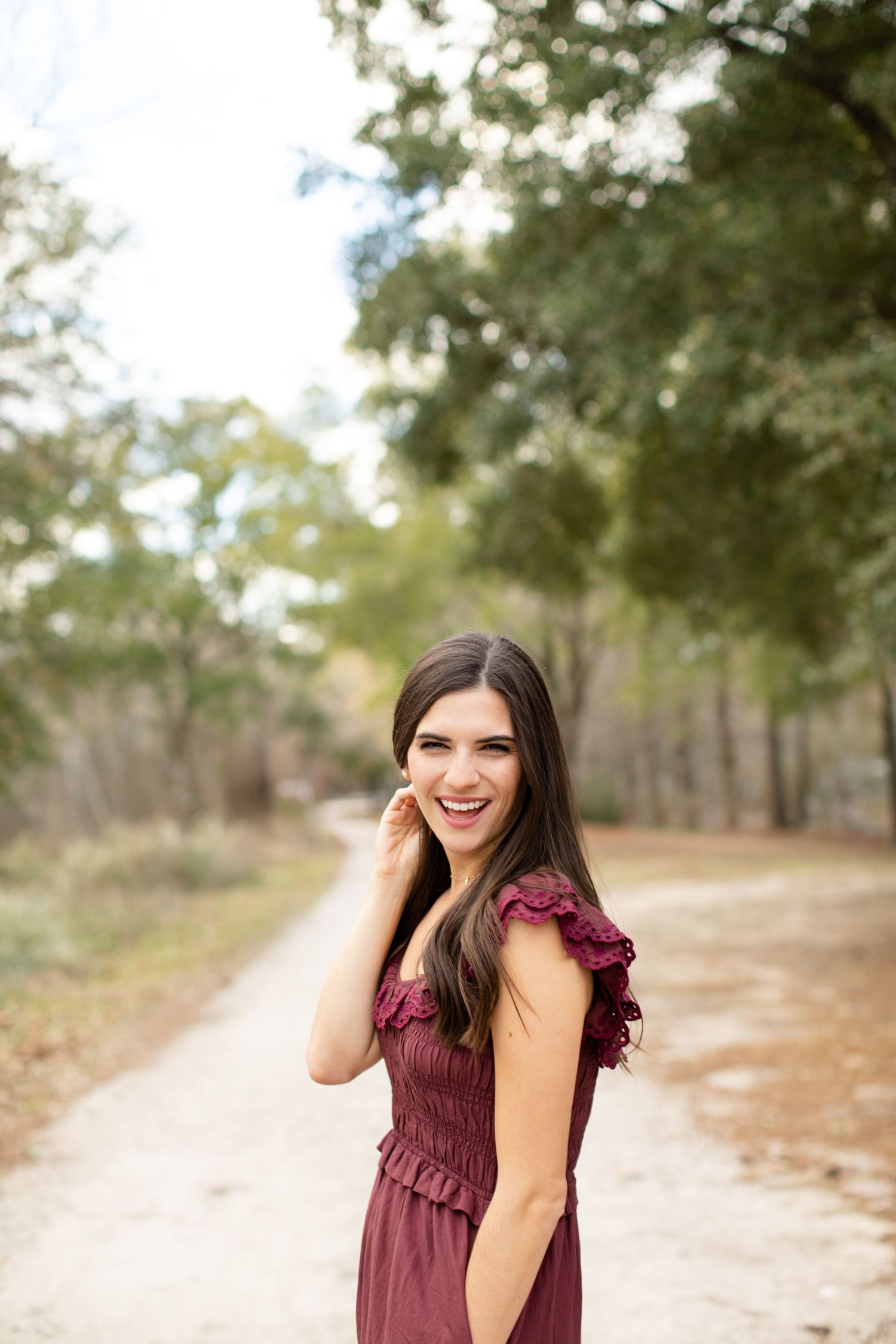 Woman in burgundy dress laughs while touching hair on nature path surrounded by trees.