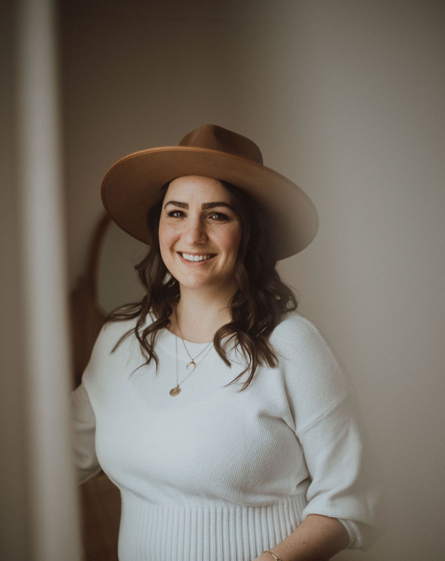 A portrait of someone wearing a brown wide-brim hat and white sweater against a neutral background.