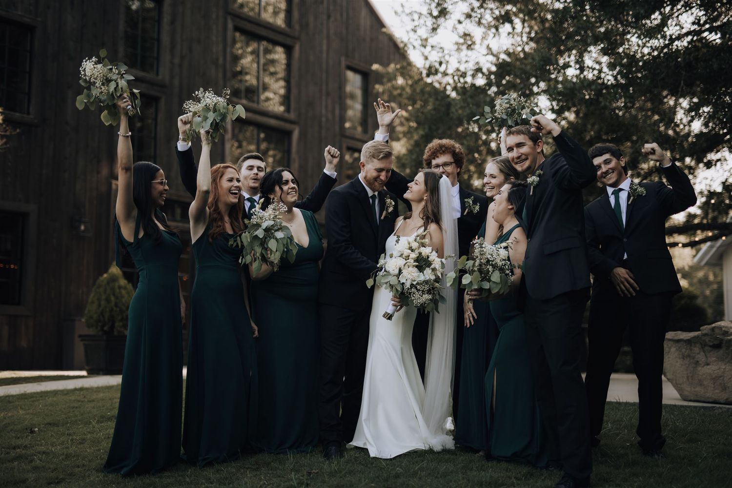 Wedding party in emerald green bridesmaid dresses and dark suits poses outdoors for group photo.