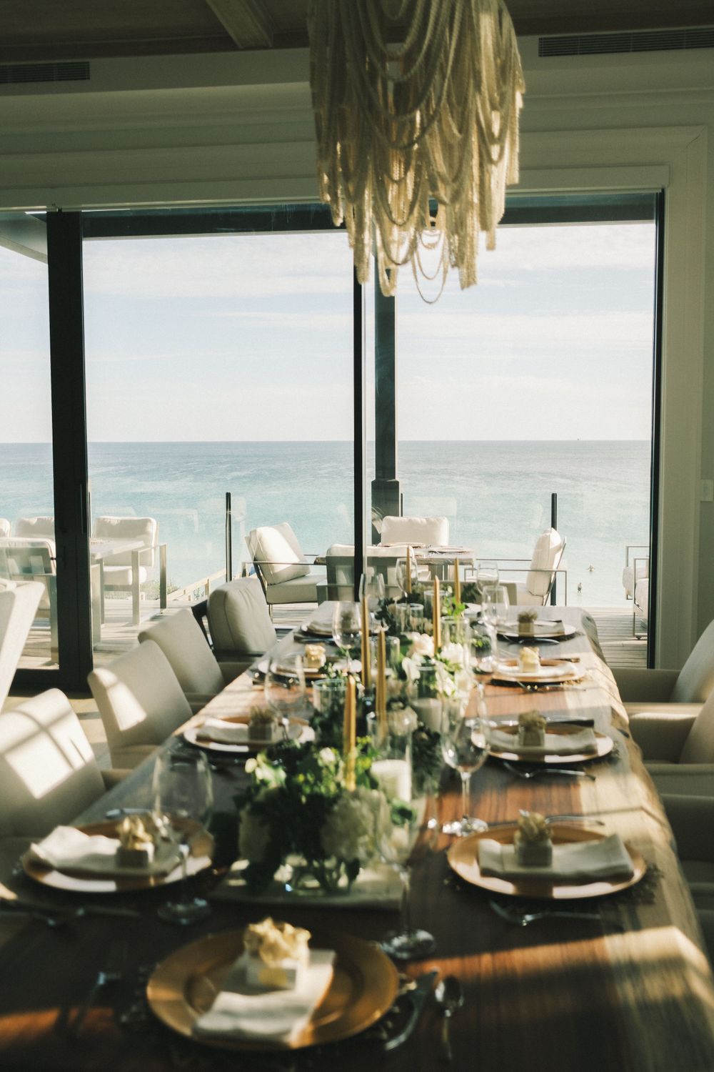 Elegant dining table setup with ocean views through large windows and natural sunlight streaming in.