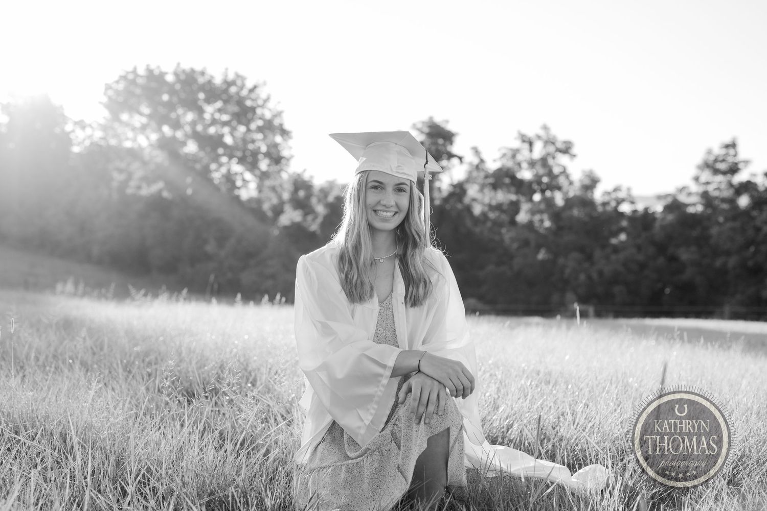 Black and white graduation photo of graduate sitting in grassy field at sunset.