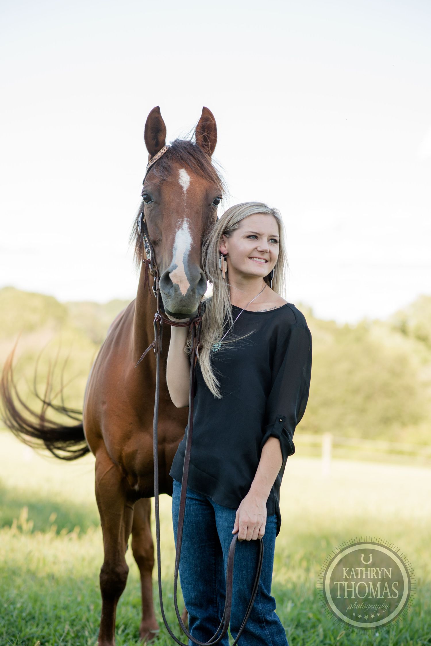 A bay horse with white blaze stands with someone in black top and jeans in field.