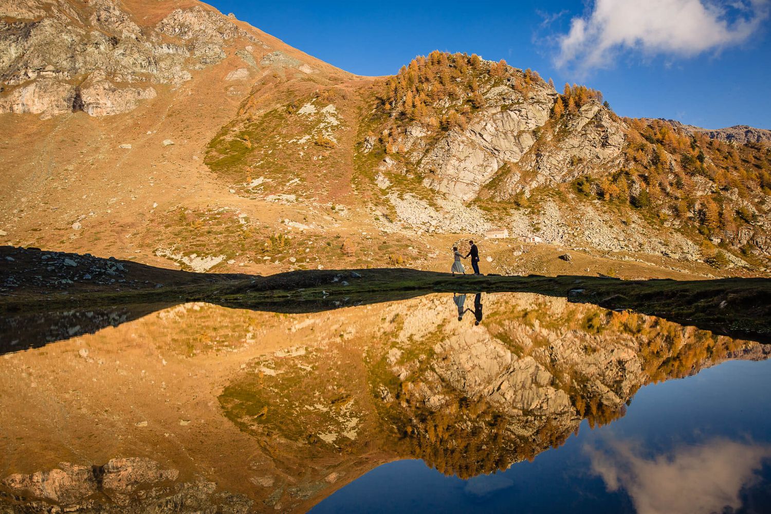 Mountain landscape perfectly reflected in alpine lake with tiny hikers visible.
