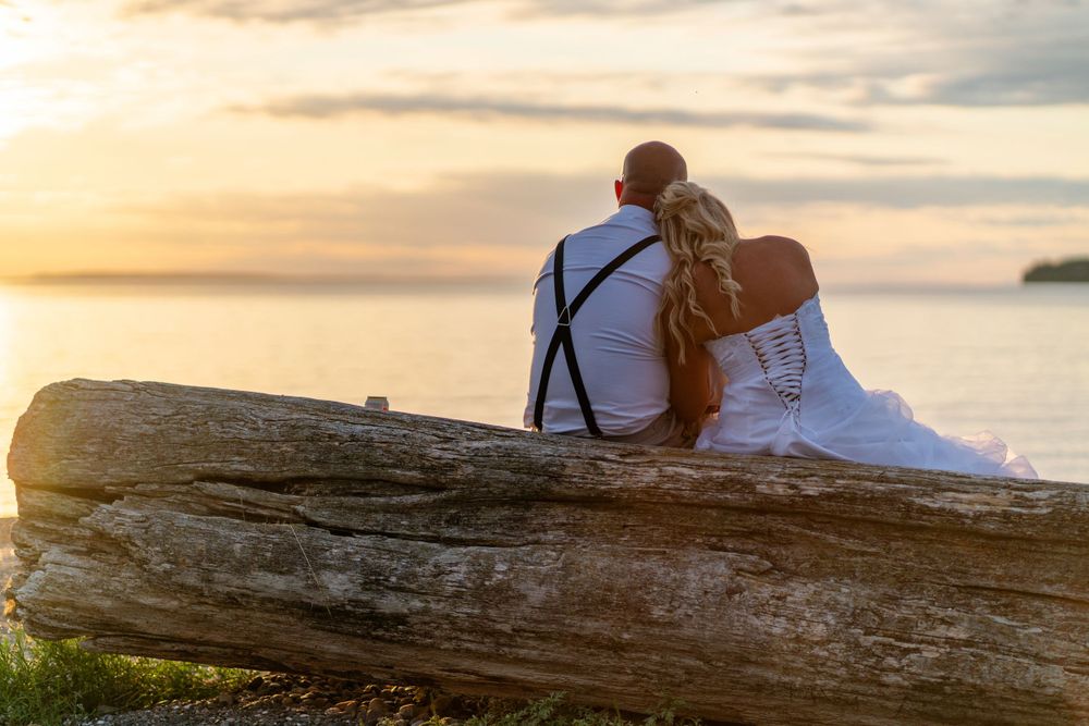 A couple sits on a driftwood log watching a golden sunset over the water.