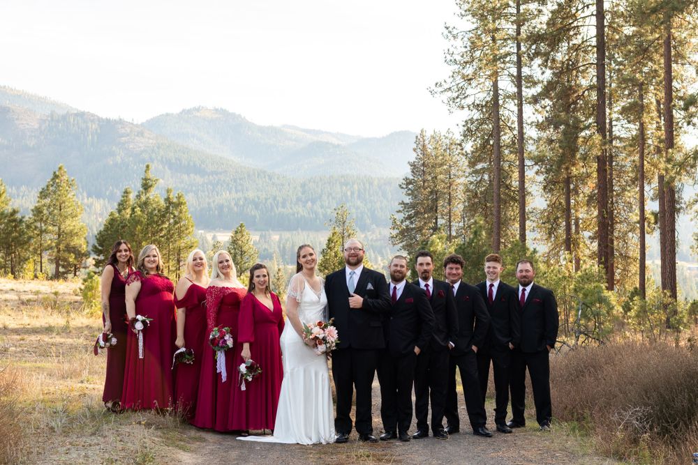 Wedding party in burgundy dresses and black suits poses on mountain path with forest backdrop.