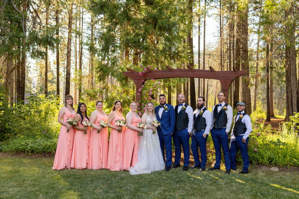 Wedding party in coral and navy attire poses under wooden arch in forest setting.