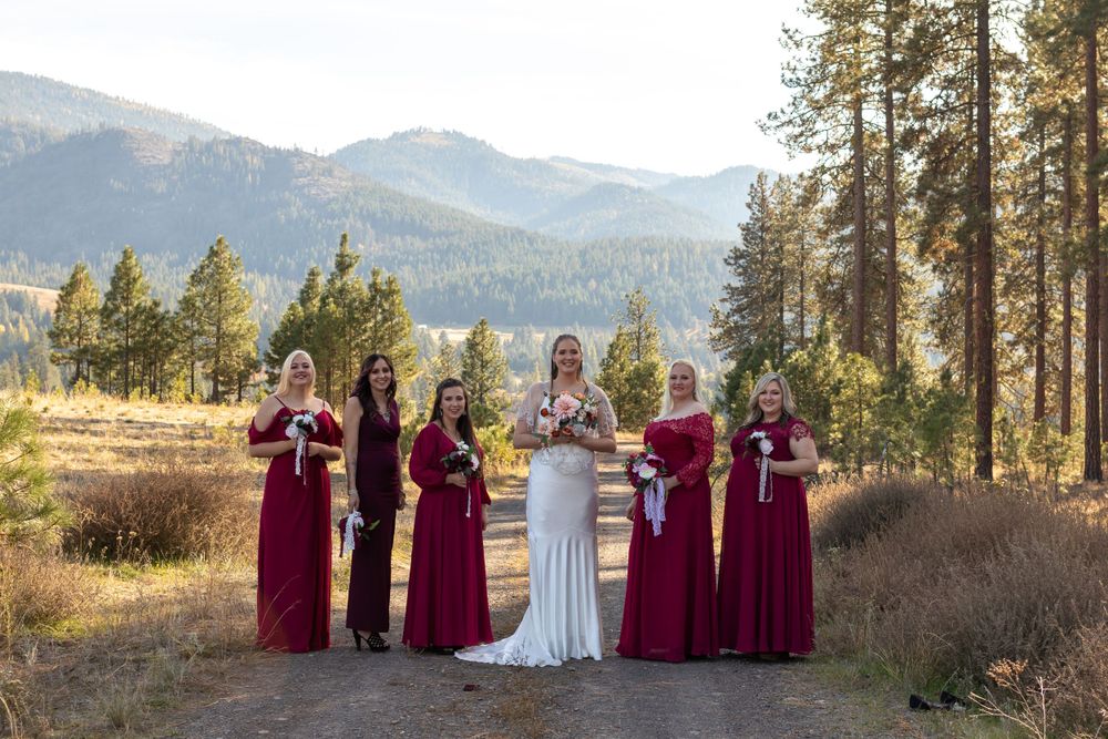 Bride in white dress stands with bridesmaids in burgundy dresses against mountain backdrop.