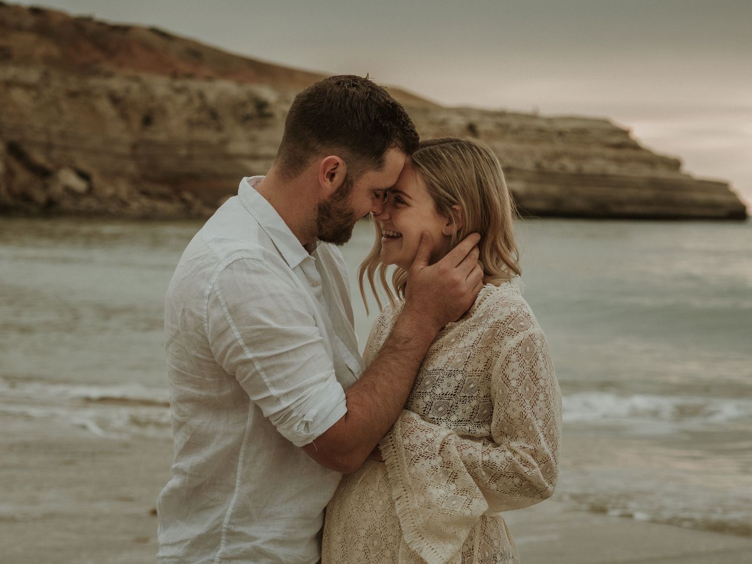 An intimate coastal moment captured between a couple in white clothing against dramatic cliffs at sunset.