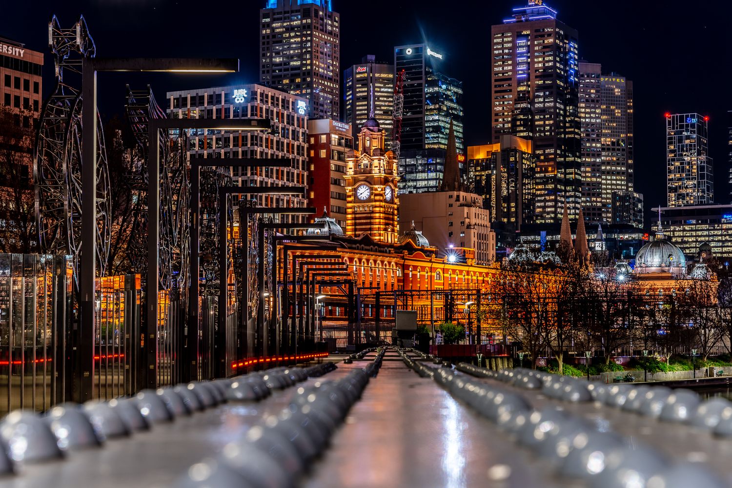 Melbourne CBD skyline at night viewed from pedestrian bridge with Flinders Street Station clock tower.