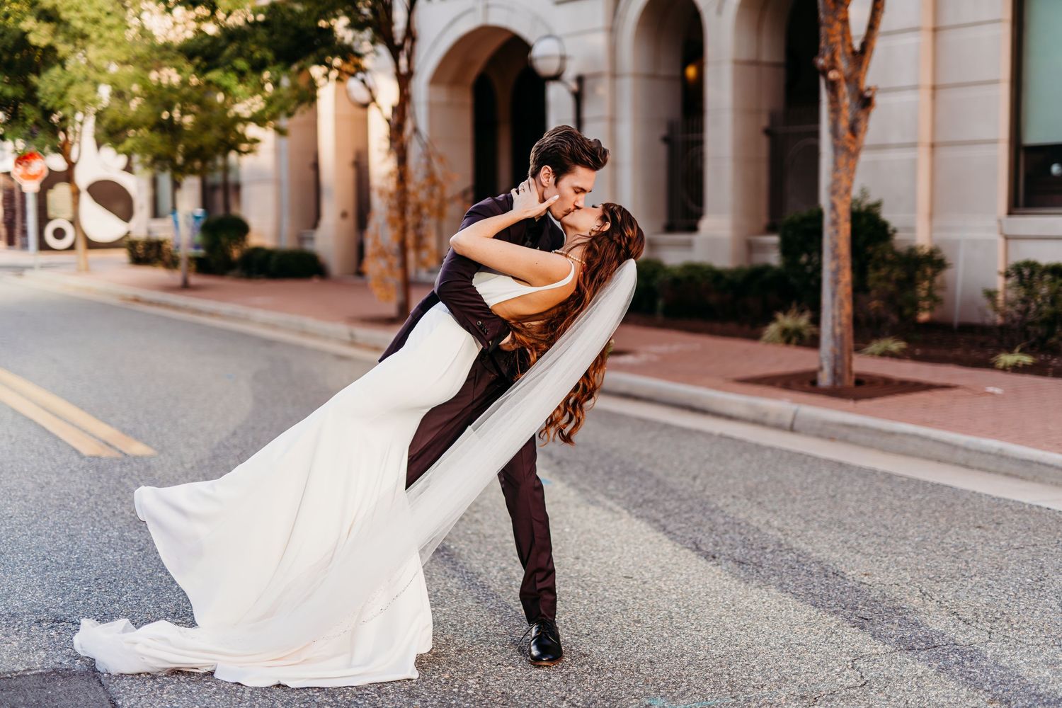 A couple performs a romantic dip pose on a sunny street with architectural details.