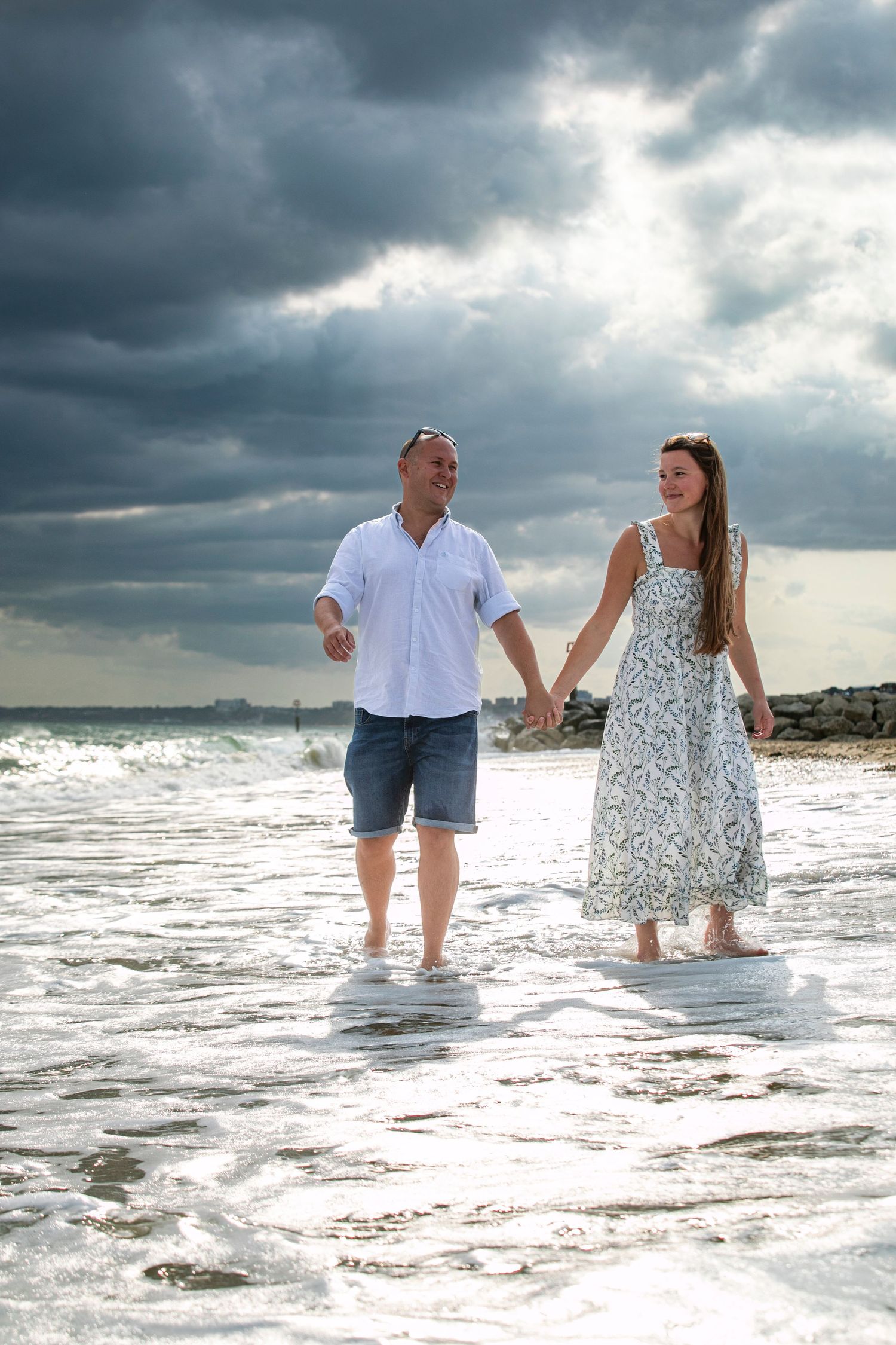 Couple wading through ocean waves at sunset with dramatic stormy clouds overhead.
