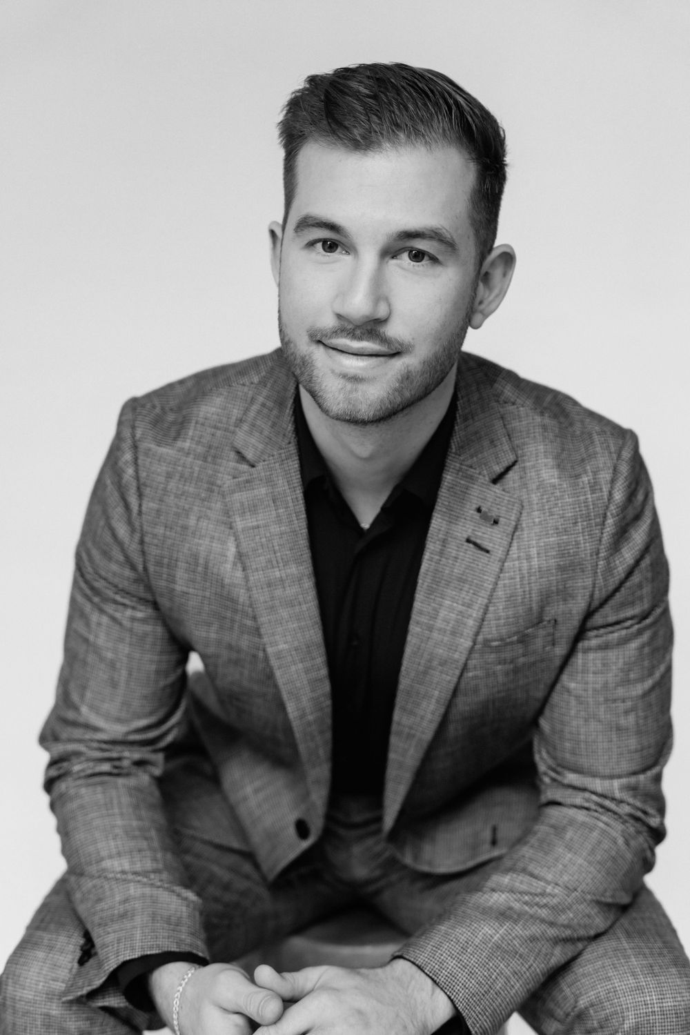 Professional headshot of person in grey suit jacket and black shirt against neutral background.