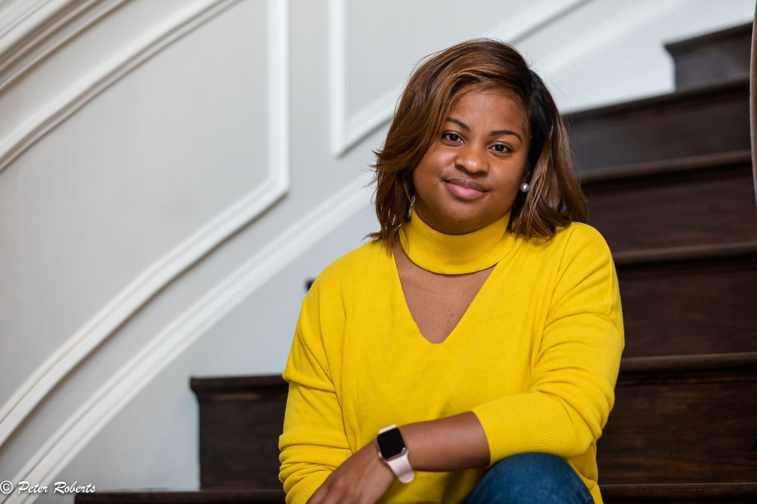 Woman in yellow turtleneck sweater sitting on wooden staircase steps in an elegant home interior.