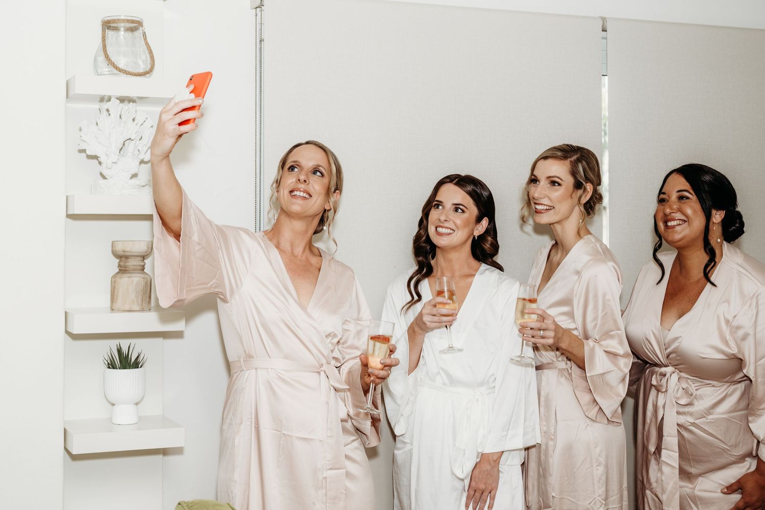 A group of women in matching silk robes take selfies while getting ready for a wedding celebration.