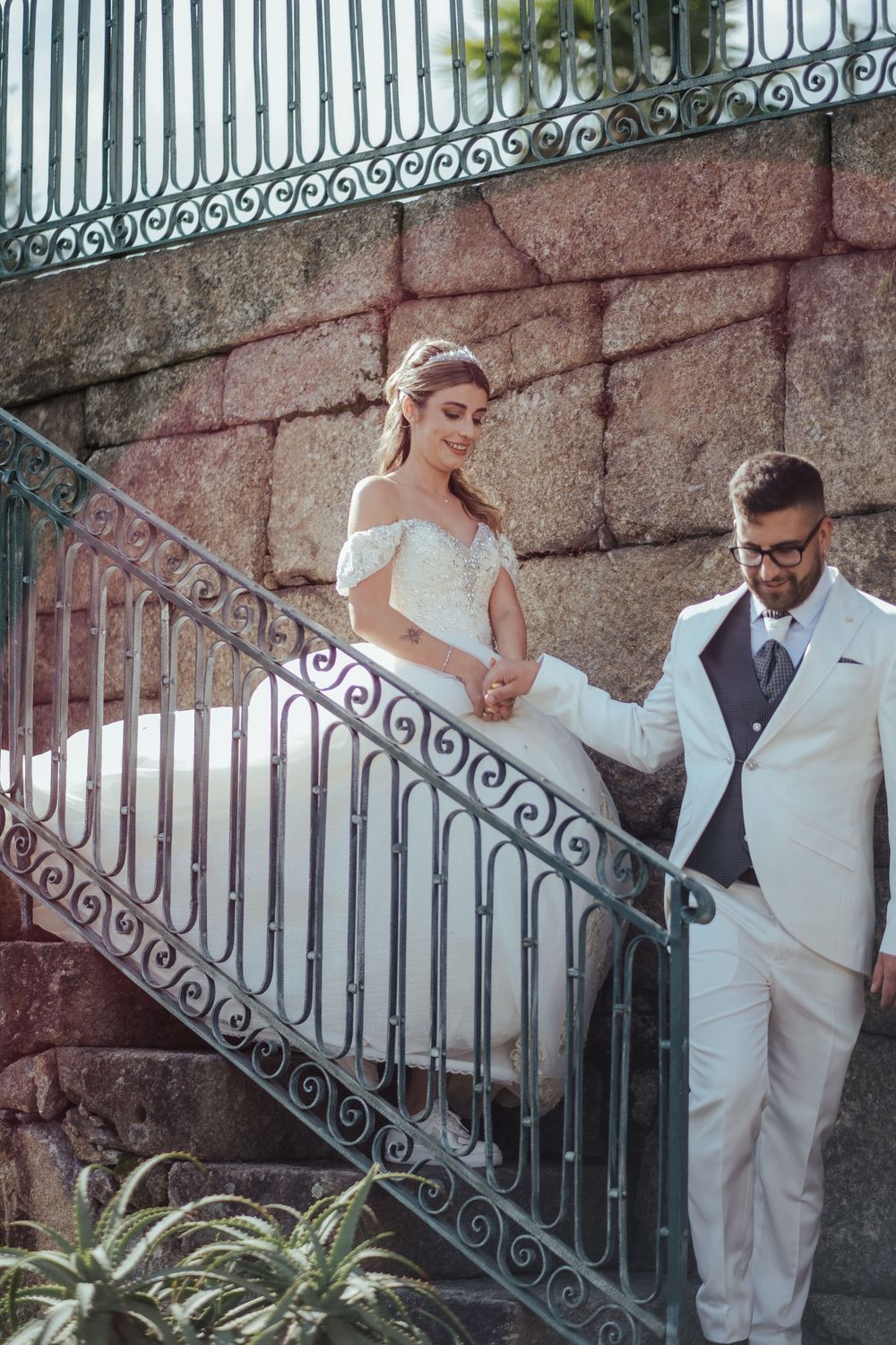Casal elegante a posar para fotos de casamento junto a grades de ferro ornamentadas e um muro de pedra.