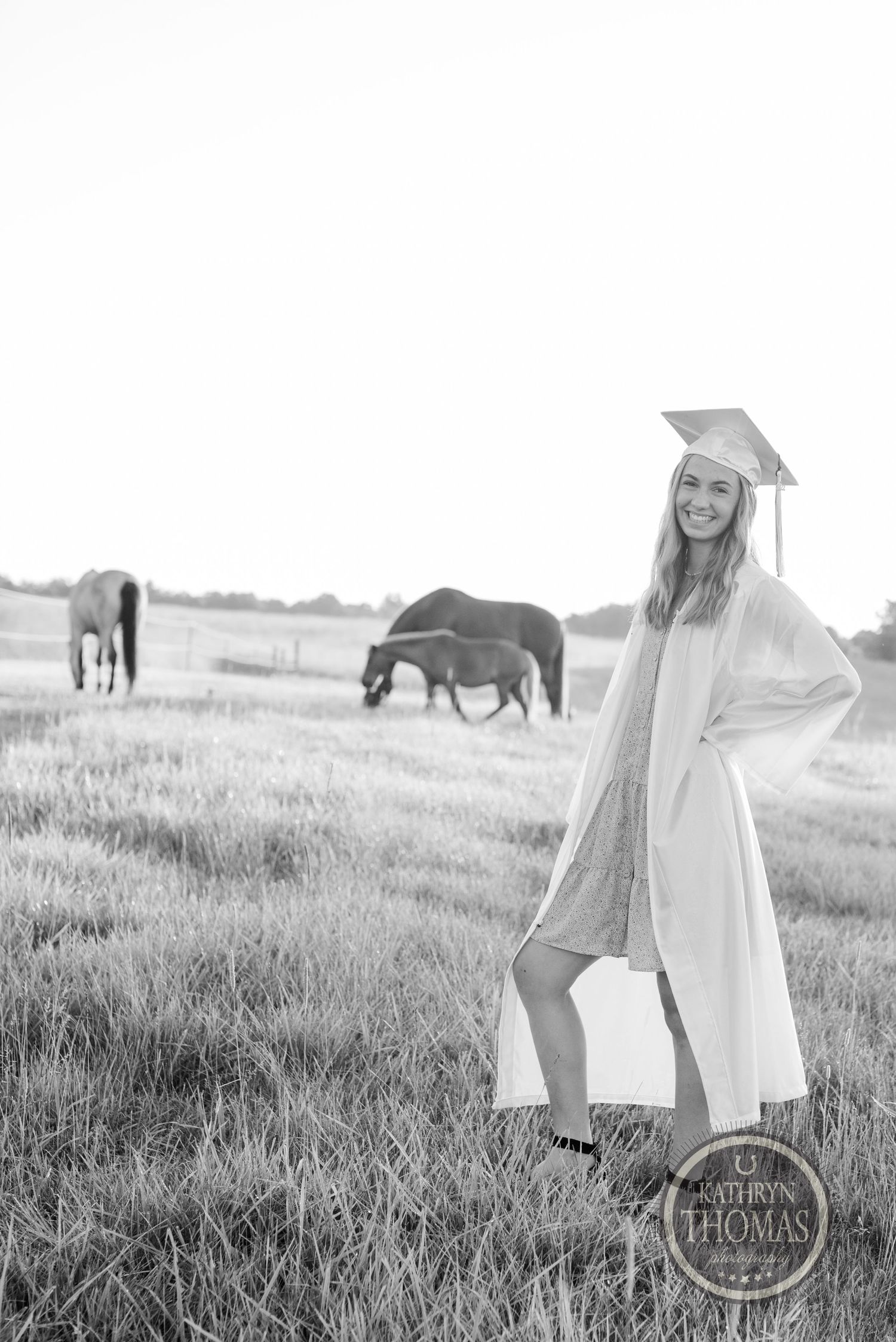 Graduate in white cap and gown poses in a sunny field with grazing horses in the background.