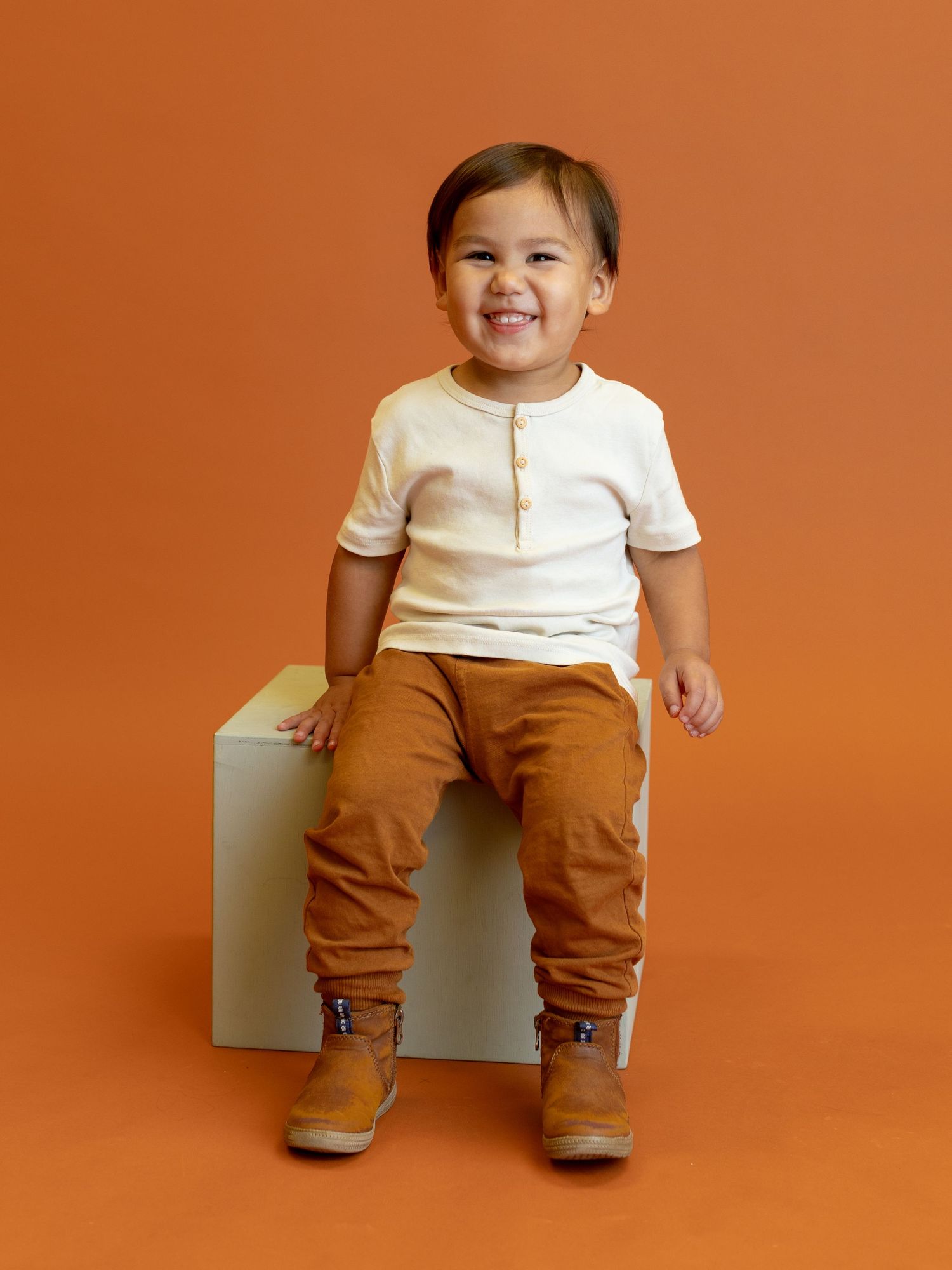 Child wearing white shirt and brown pants sitting on white cube against orange background.