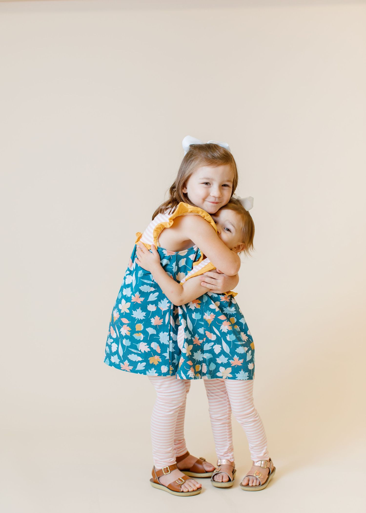 Two children in matching blue floral dresses sharing a warm hug against a light background.
