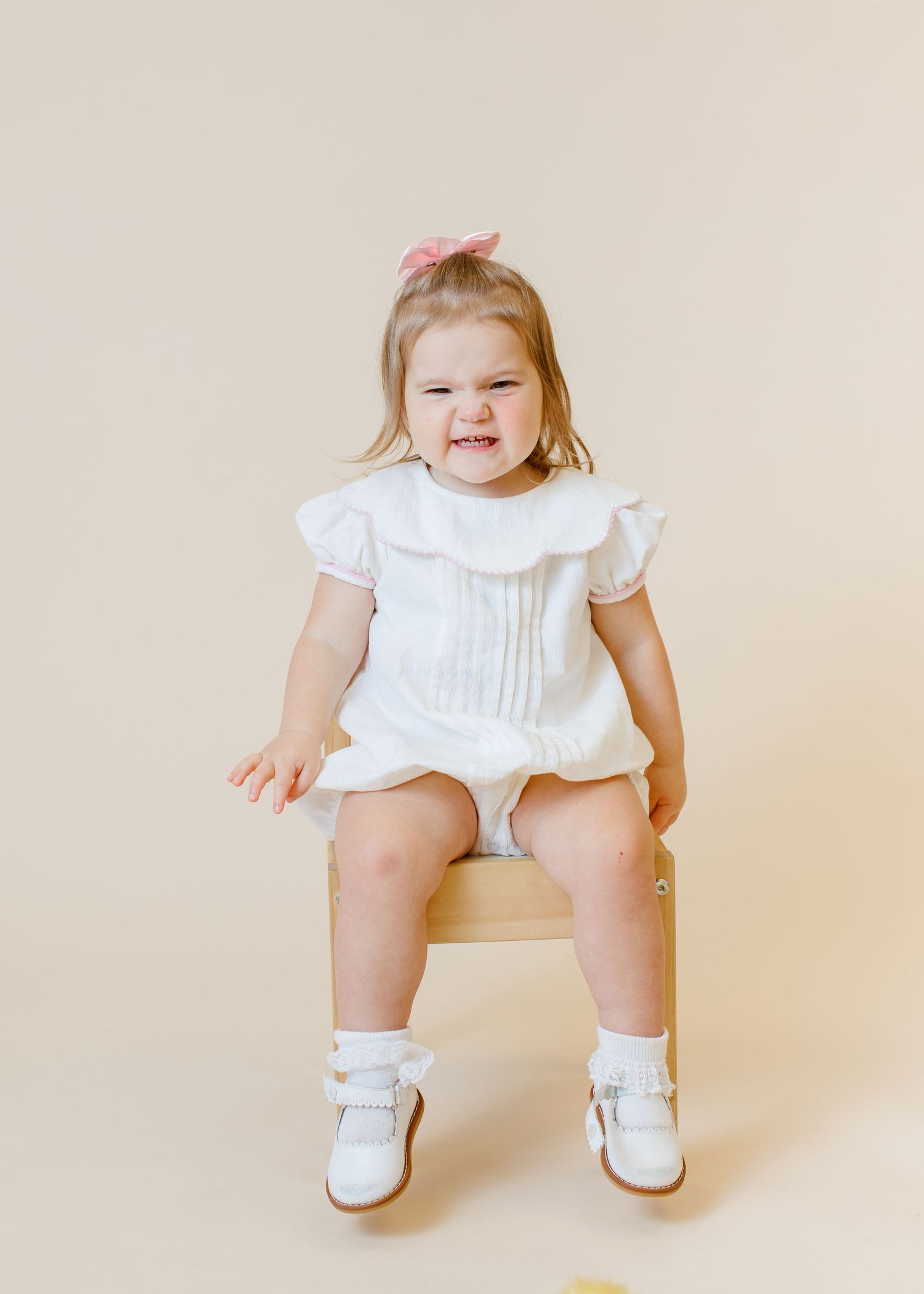 Child in white ruffled dress sitting on wooden chair with joyful expression.