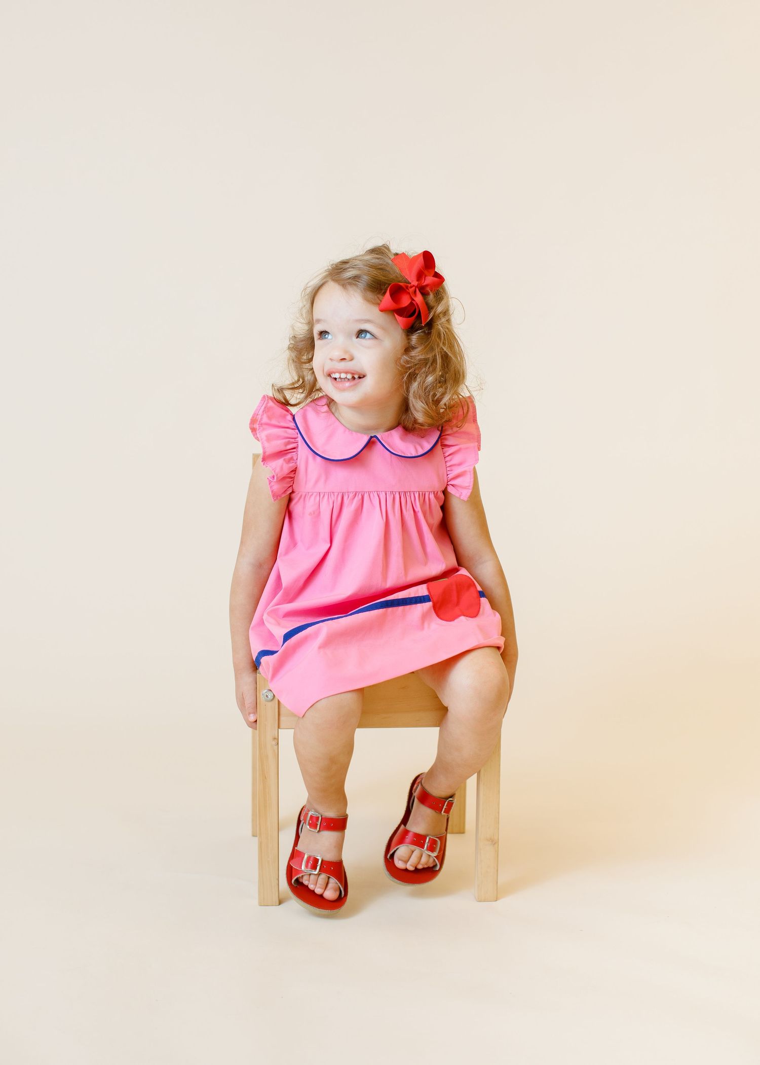 Child in pink dress with red flower accessory sits cheerfully on wooden chair.