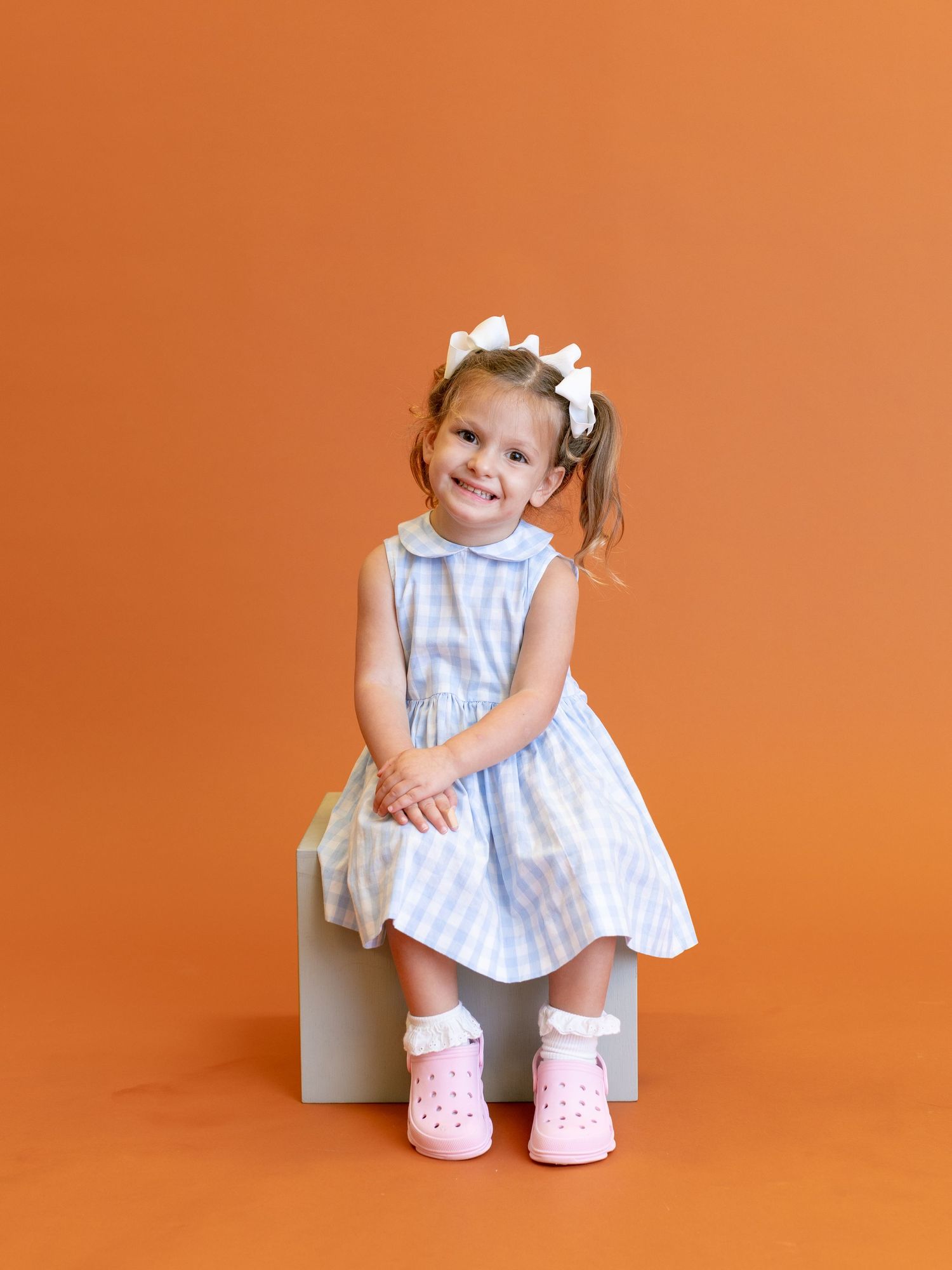 Child in white gingham dress and pink shoes sitting on cube against orange background.