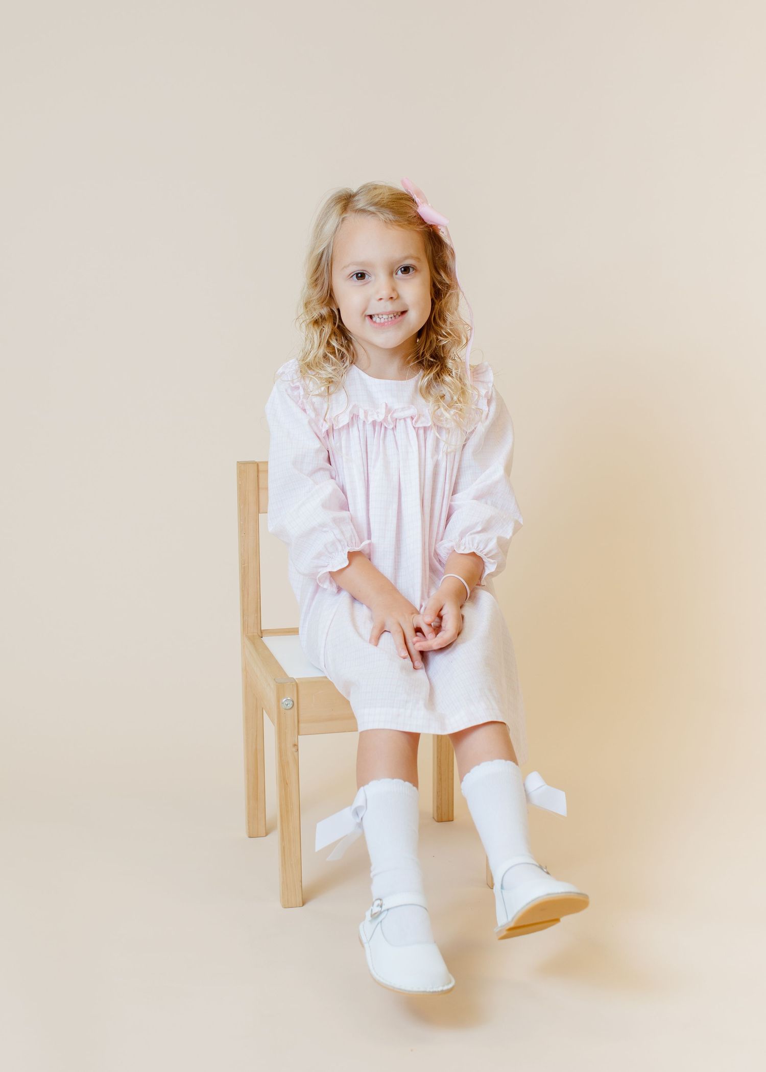 Child in white smocked dress with ruffles sitting prettily on wooden chair.