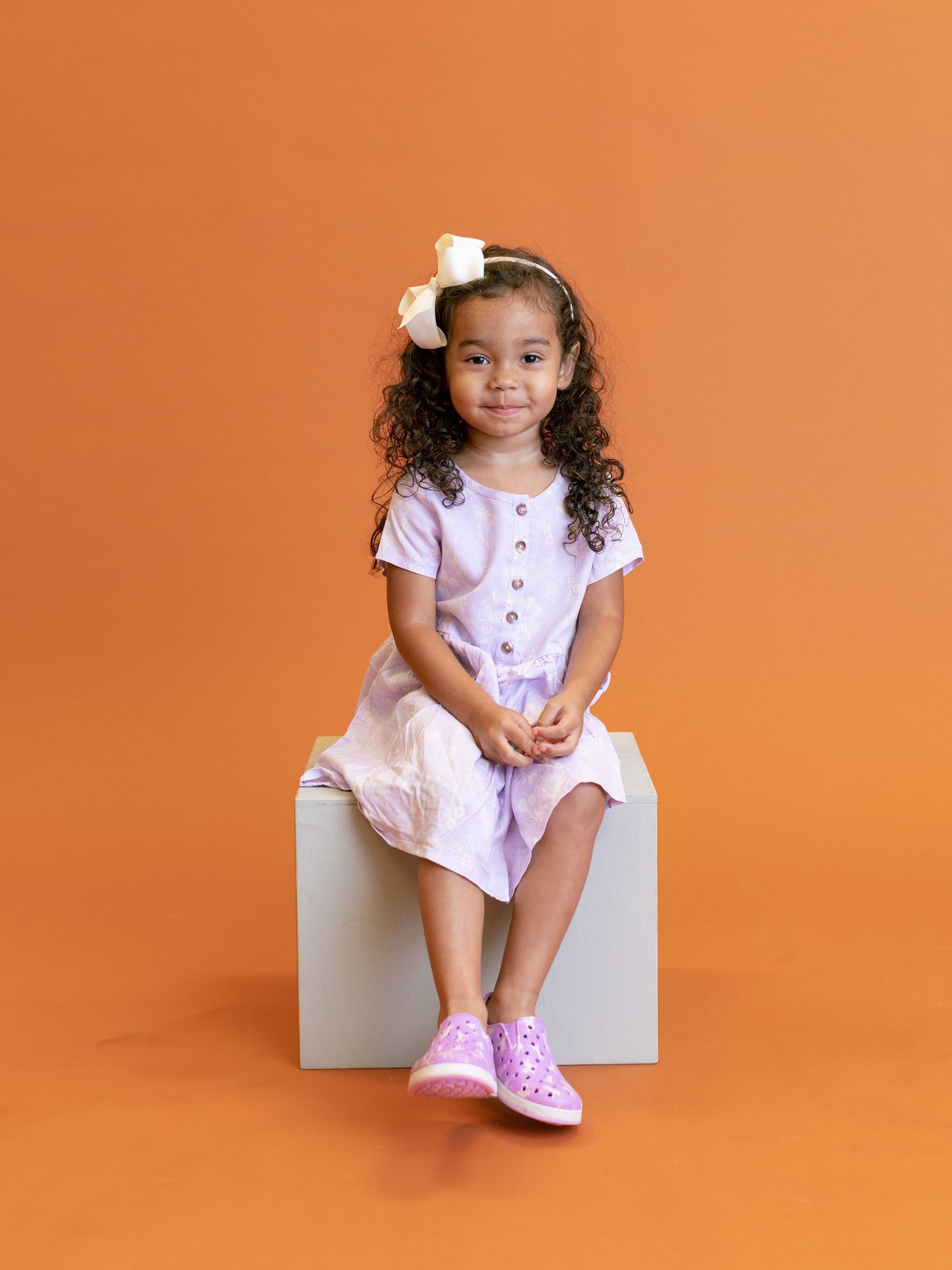 Young child in pink dress and matching shoes sitting on white cube against orange backdrop.