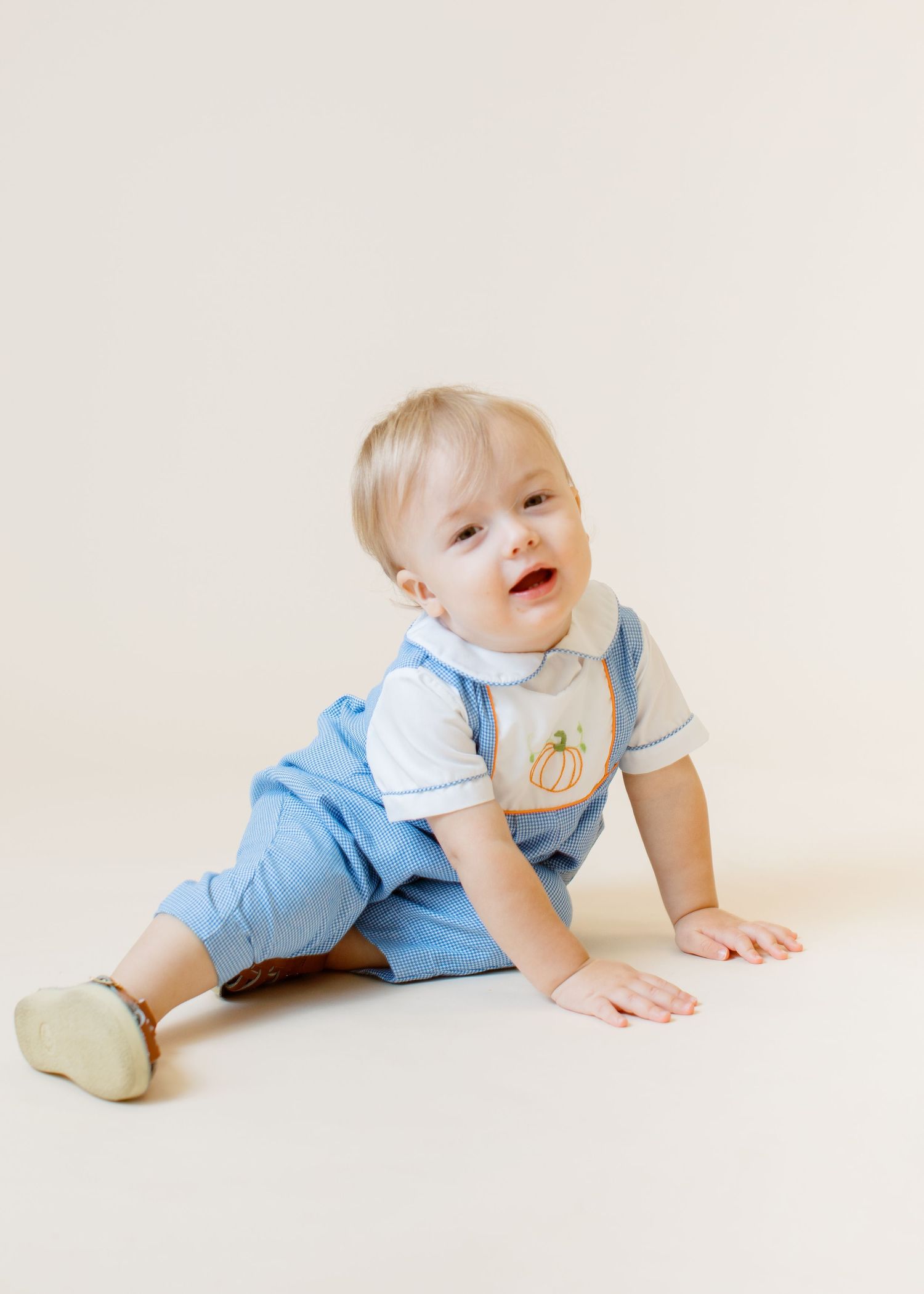 Infant in light blue outfit with white trim sits on floor in playful pose against neutral backdrop.