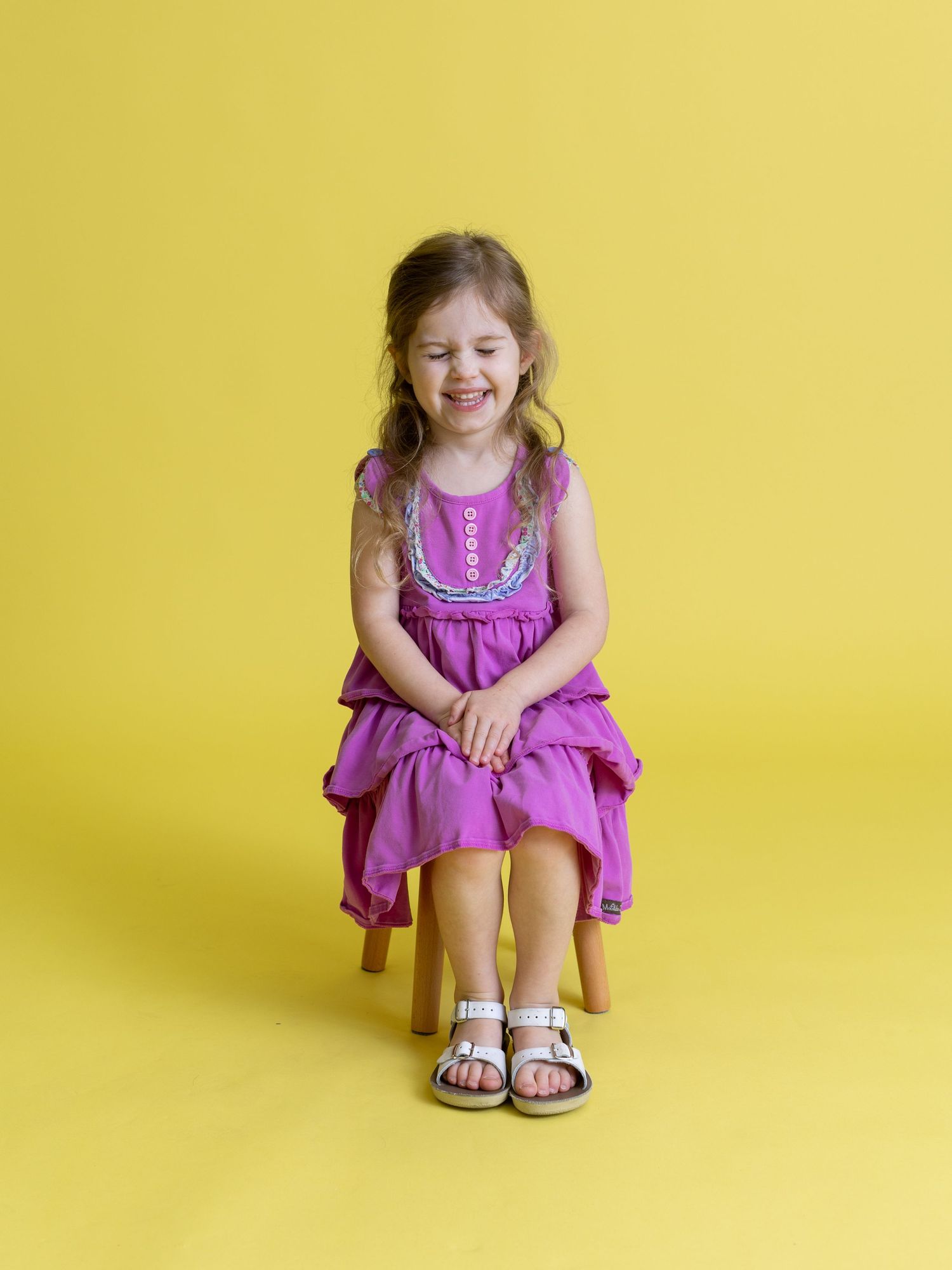 Child in purple dress sitting on wooden stool against yellow background.