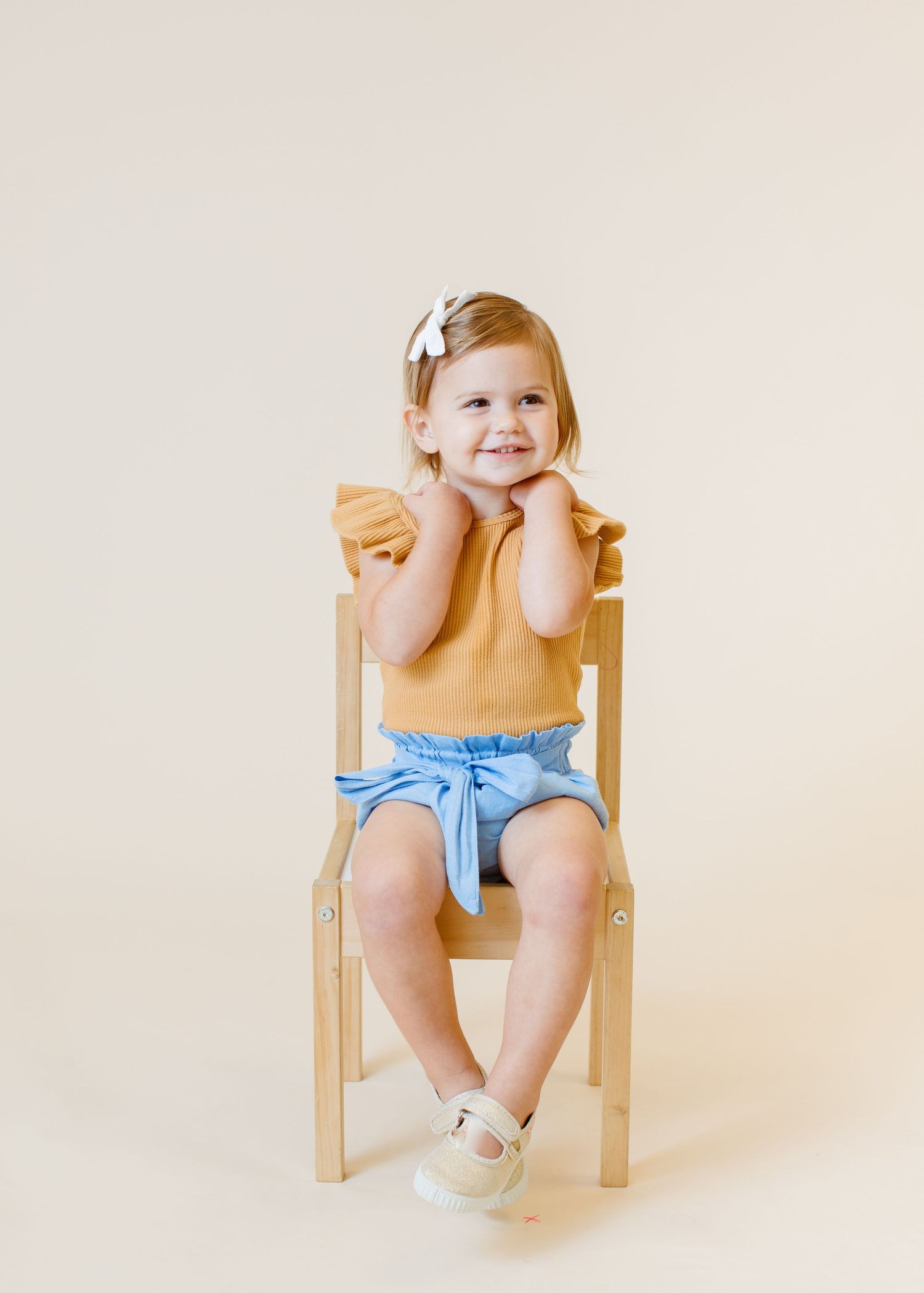 Child in yellow top and blue shorts sits casually on wooden chair with sweet smile.