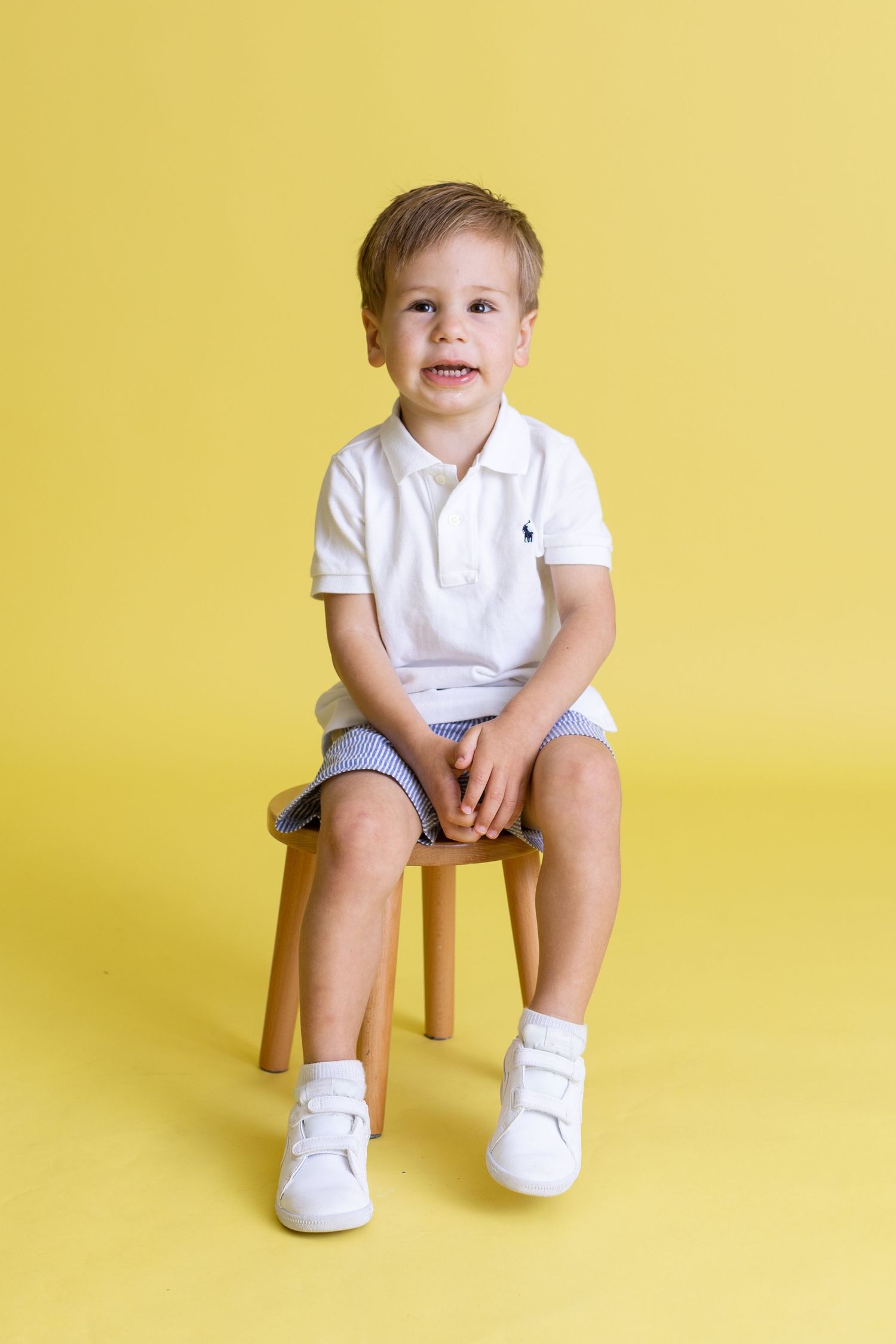 Child in white polo and shorts sitting on wooden stool against yellow background.