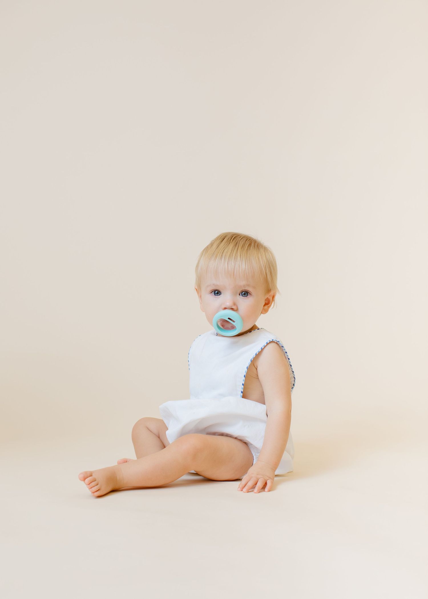 Baby in white sleeveless romper with blue pacifier sits on neutral background.