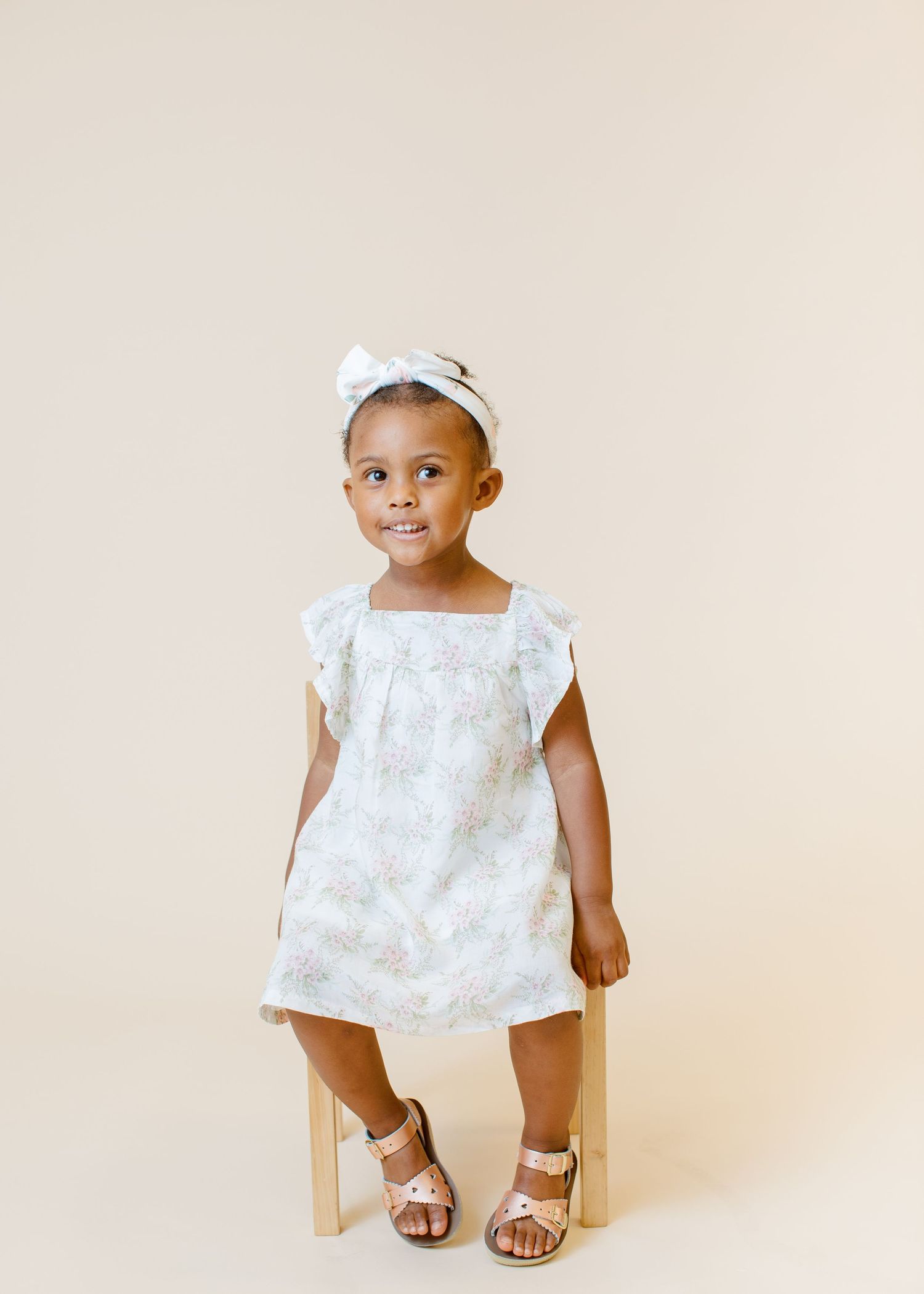Sweet portrait of a toddler in white dress and bow sitting on wooden chair against neutral backdrop.